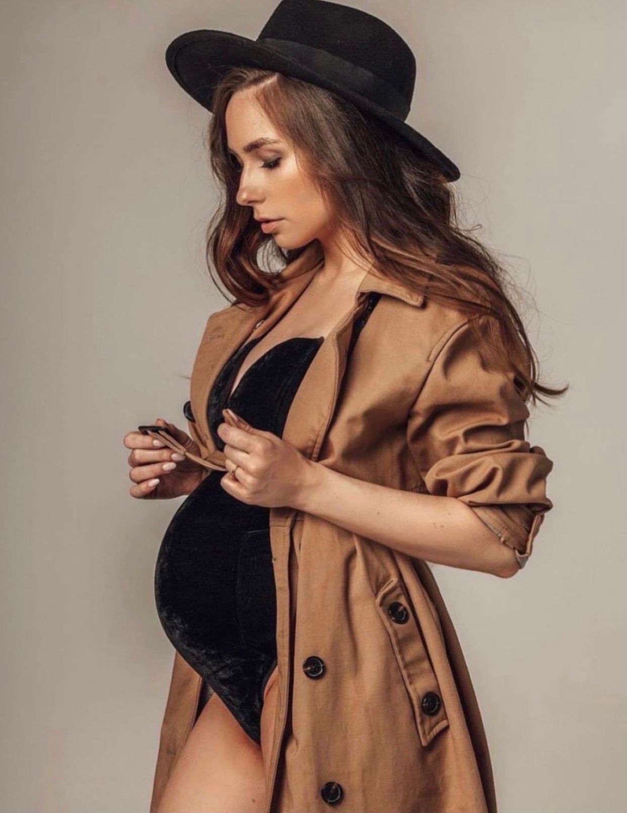Pregnant woman in black bodysuit, camel trench coat, and black fedora hat poses elegantly in a maternity fashion shoot.