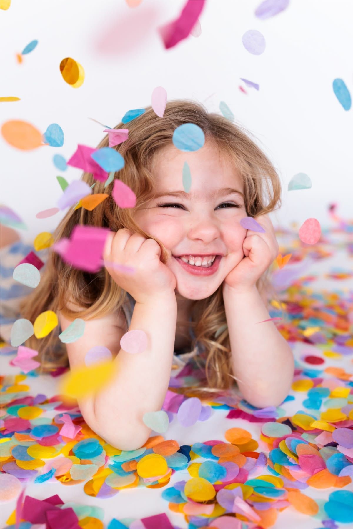 Joyful little girl laughing while lying in colorful confetti falling around her.