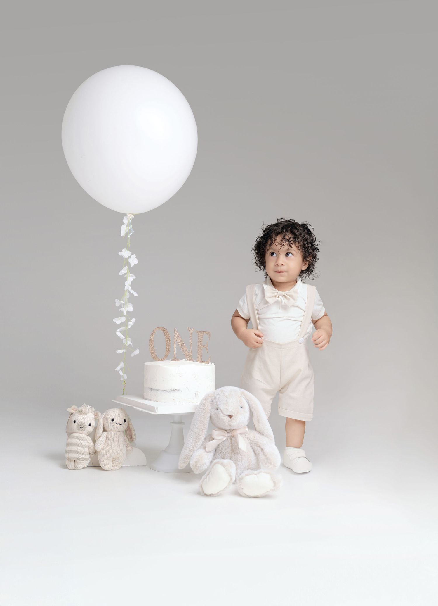 Toddler in beige outfit stands beside first birthday cake, white balloon, and stuffed bunny toys.