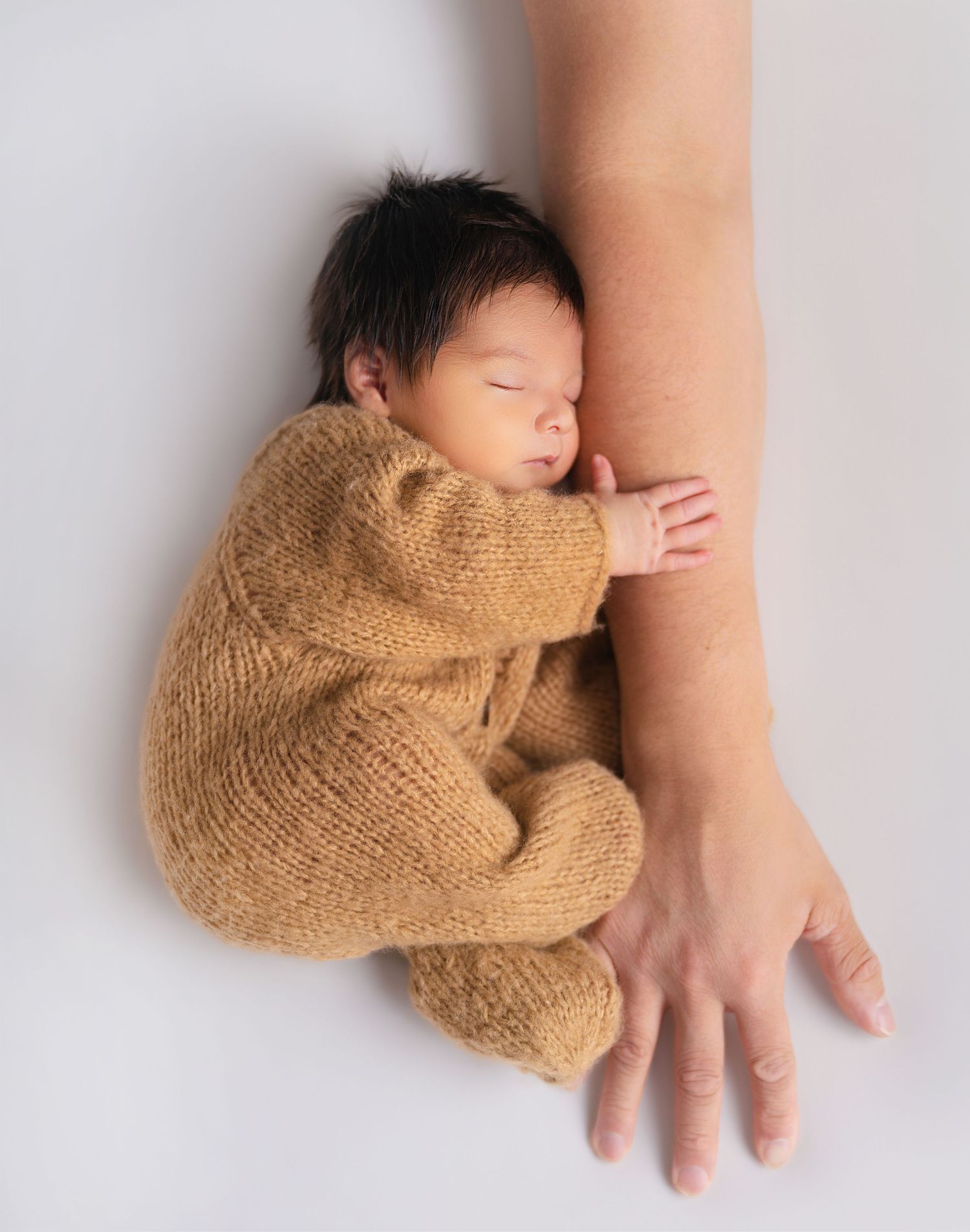 Newborn baby in tan knit onesie sleeping peacefully beside an adult's arm on white background.