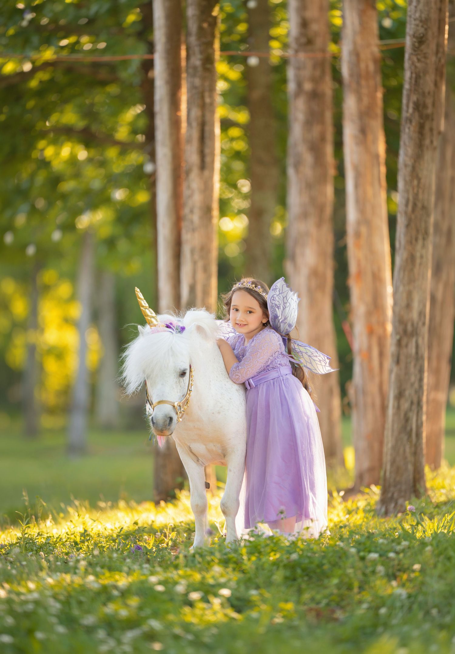 A young girl in a purple fairy dress hugs a white unicorn pony in a sunlit forest clearing.