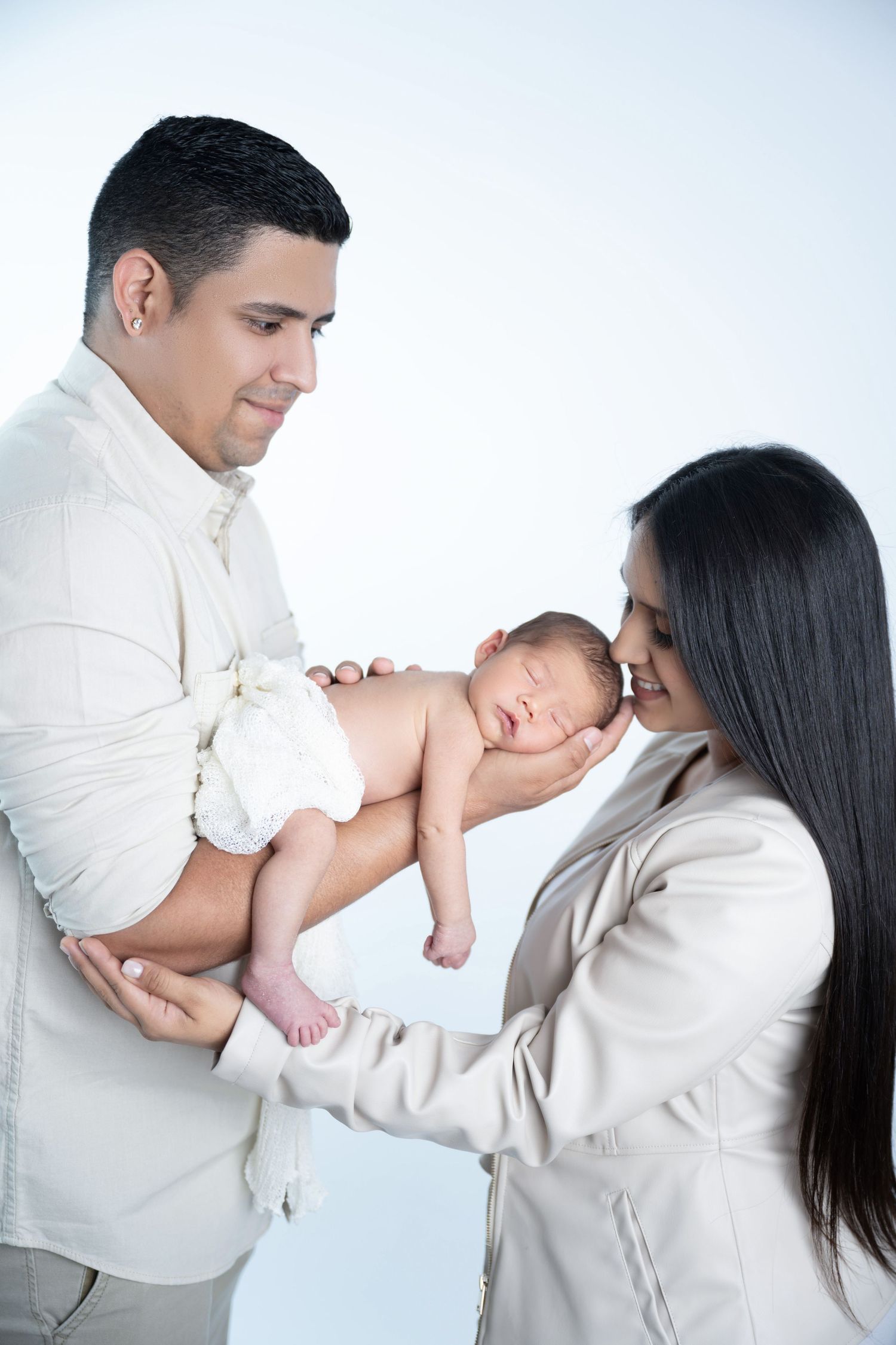 Parents in white outfits holding sleeping newborn against white backdrop.