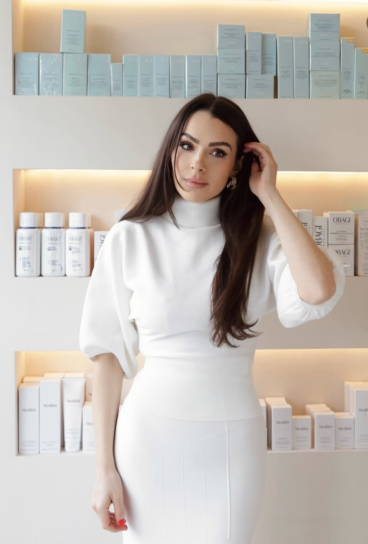 Woman in white dress poses in front of shelves displaying Obagi skincare products in a modern clinic.