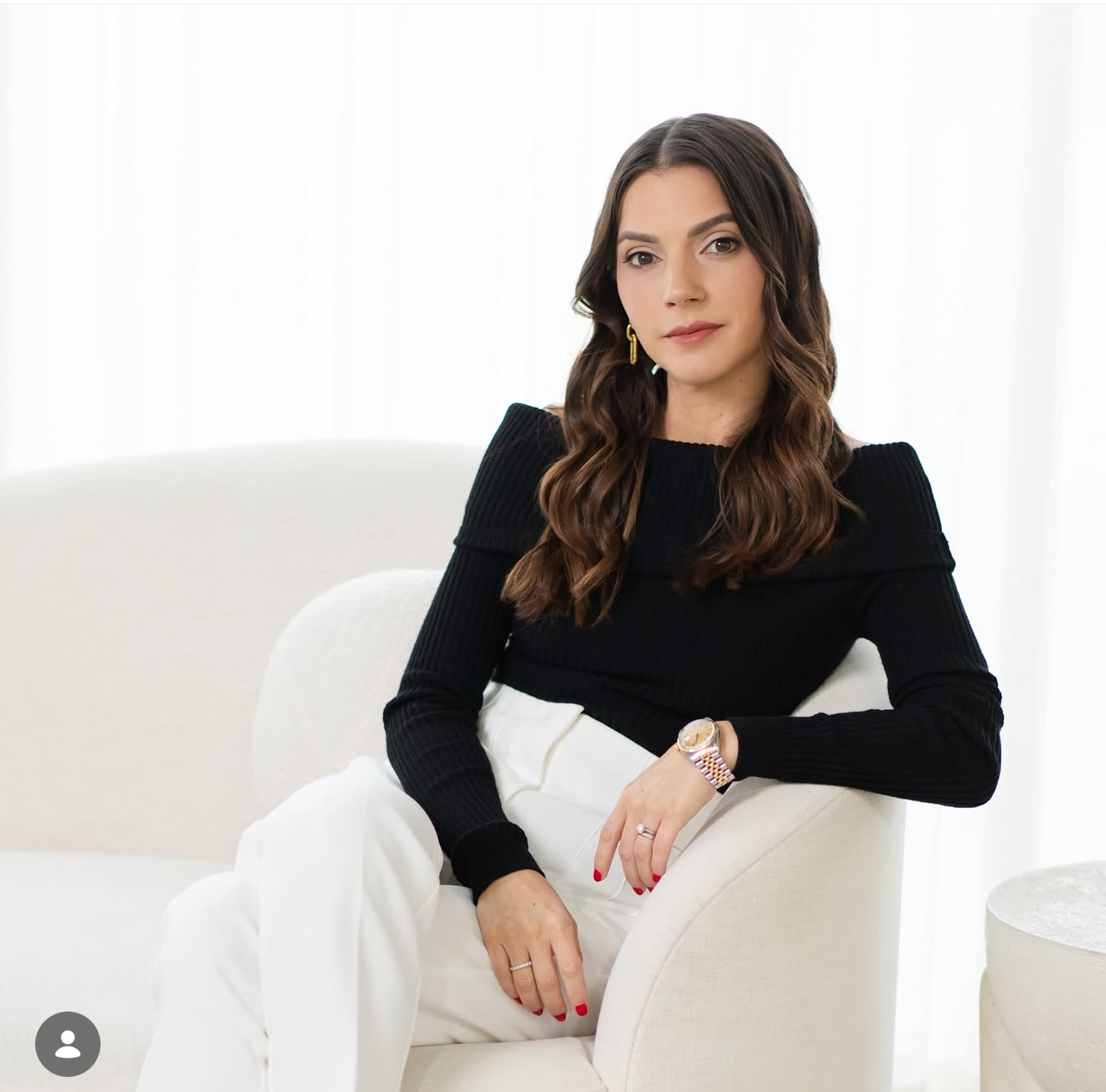 Woman in black top and white pants sitting elegantly in a white chair, wearing a gold ring and watch.