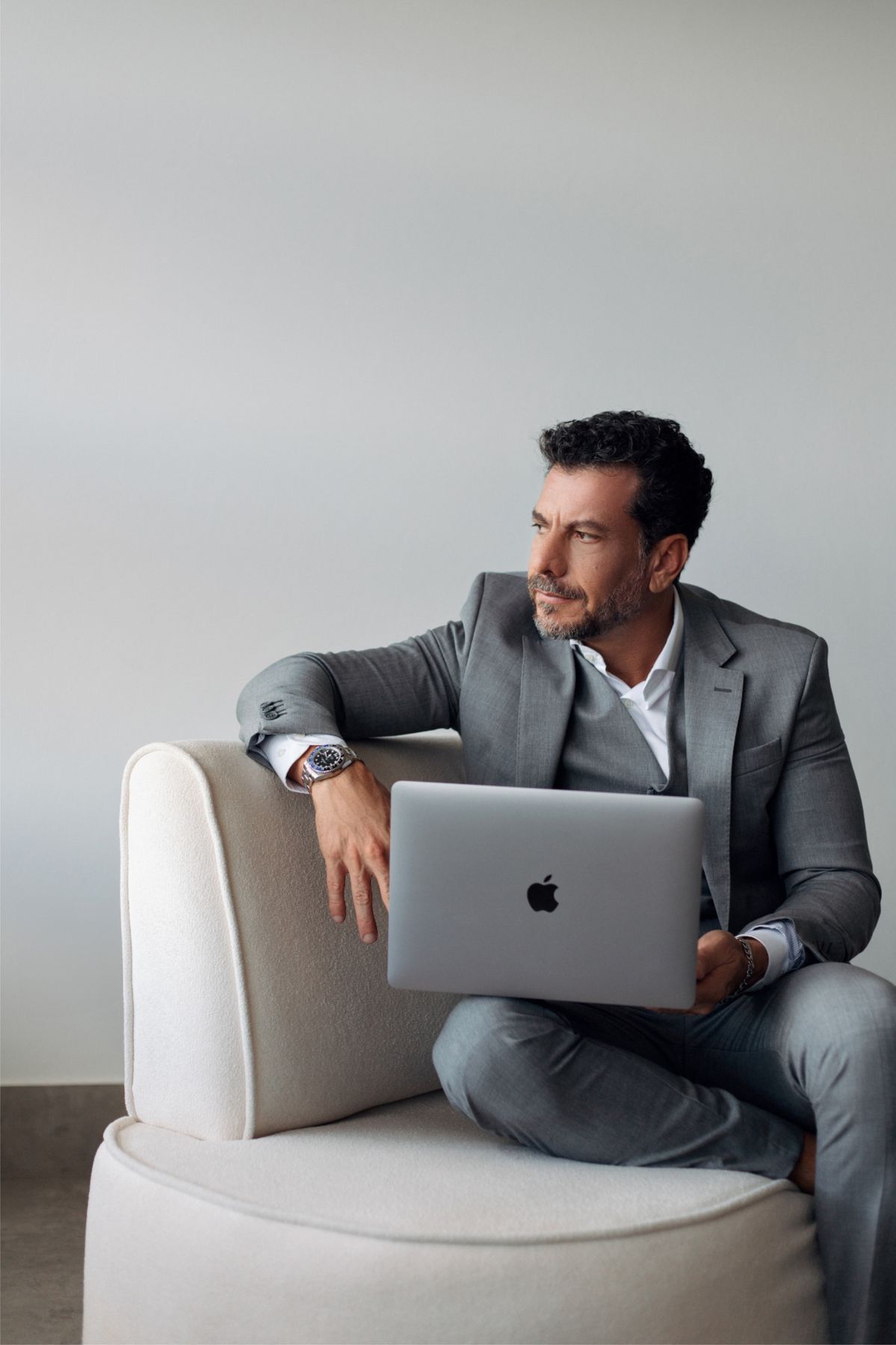 Professional man in gray suit sitting in white chair with Apple laptop, wearing luxury watch, looking away thoughtfully.