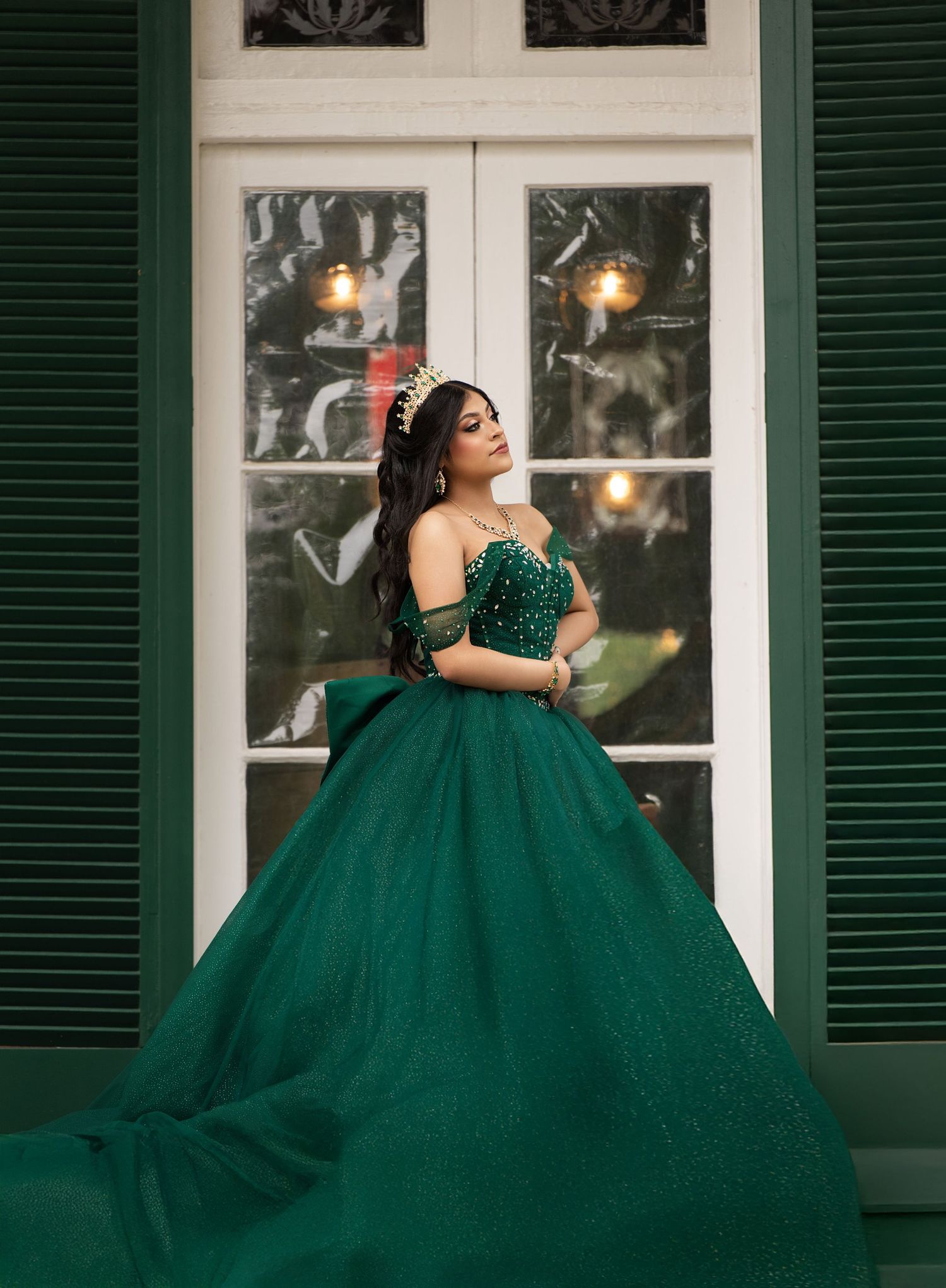 Young woman in emerald green ball gown with gold tiara poses elegantly before white-trimmed doors.