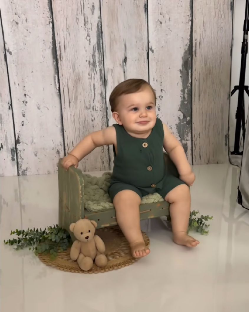 A small child sits on a rustic wooden box wearing a green romper, with a teddy bear placed beside them.