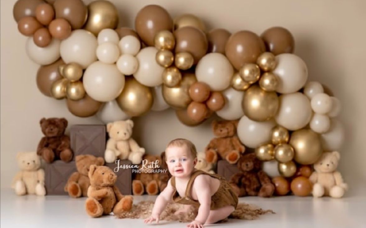 Baby surrounded by brown and gold balloons with teddy bears for a milestone photo session.