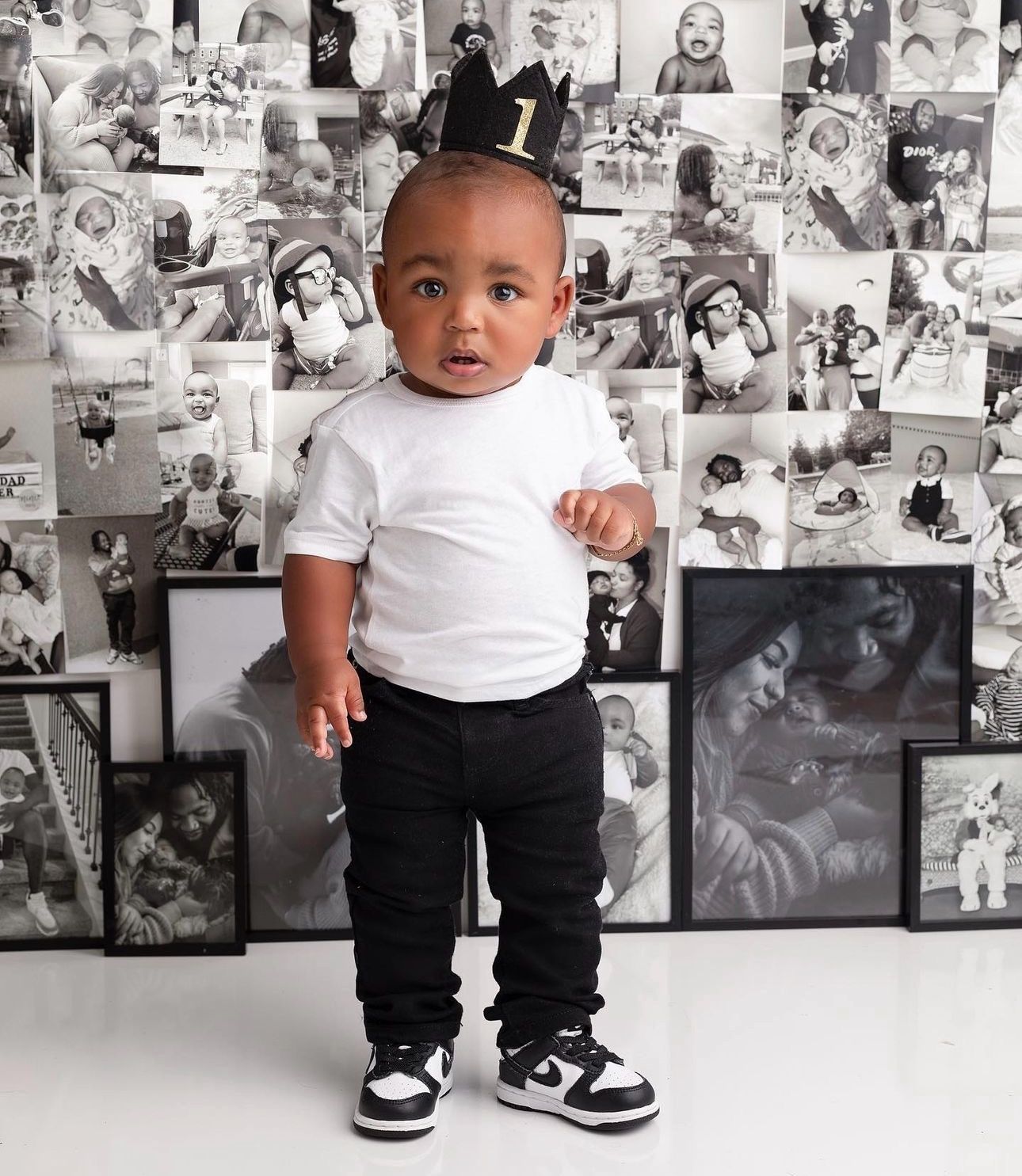 Small child in white t-shirt and black pants standing against wall covered in black and white photographs.