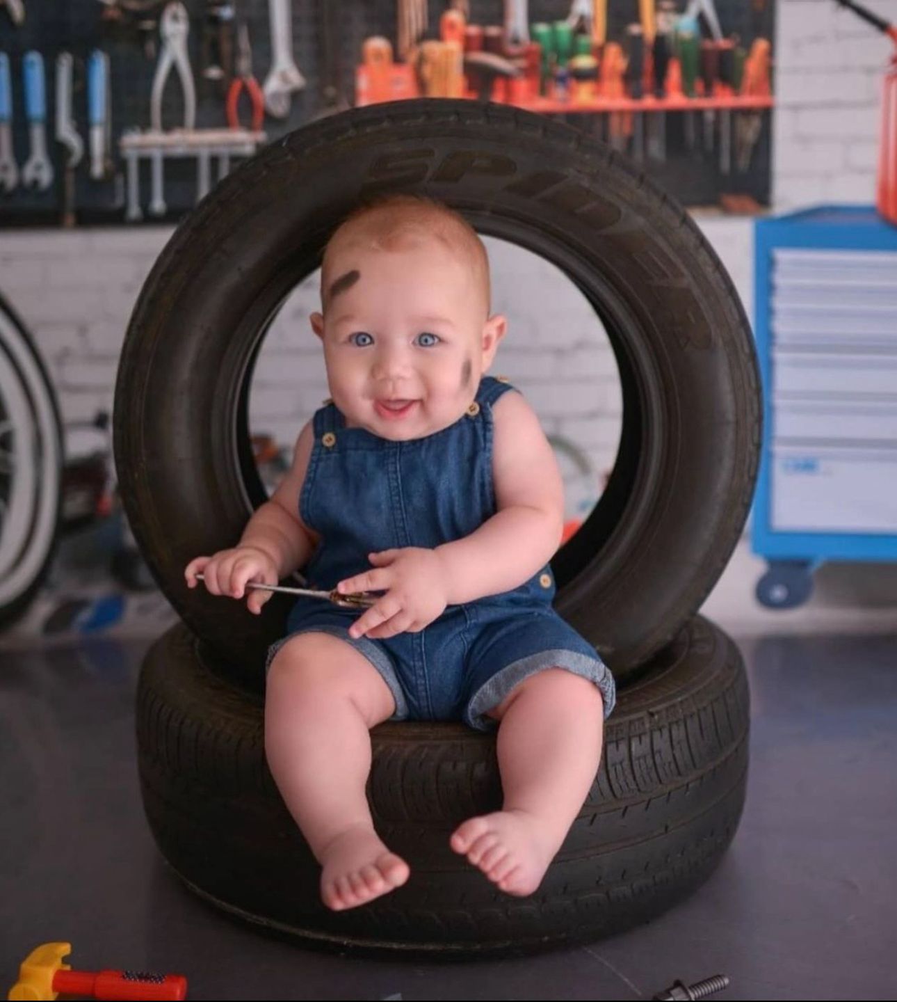 Baby in blue overalls sits nestled inside stacked tires at an auto repair shop.