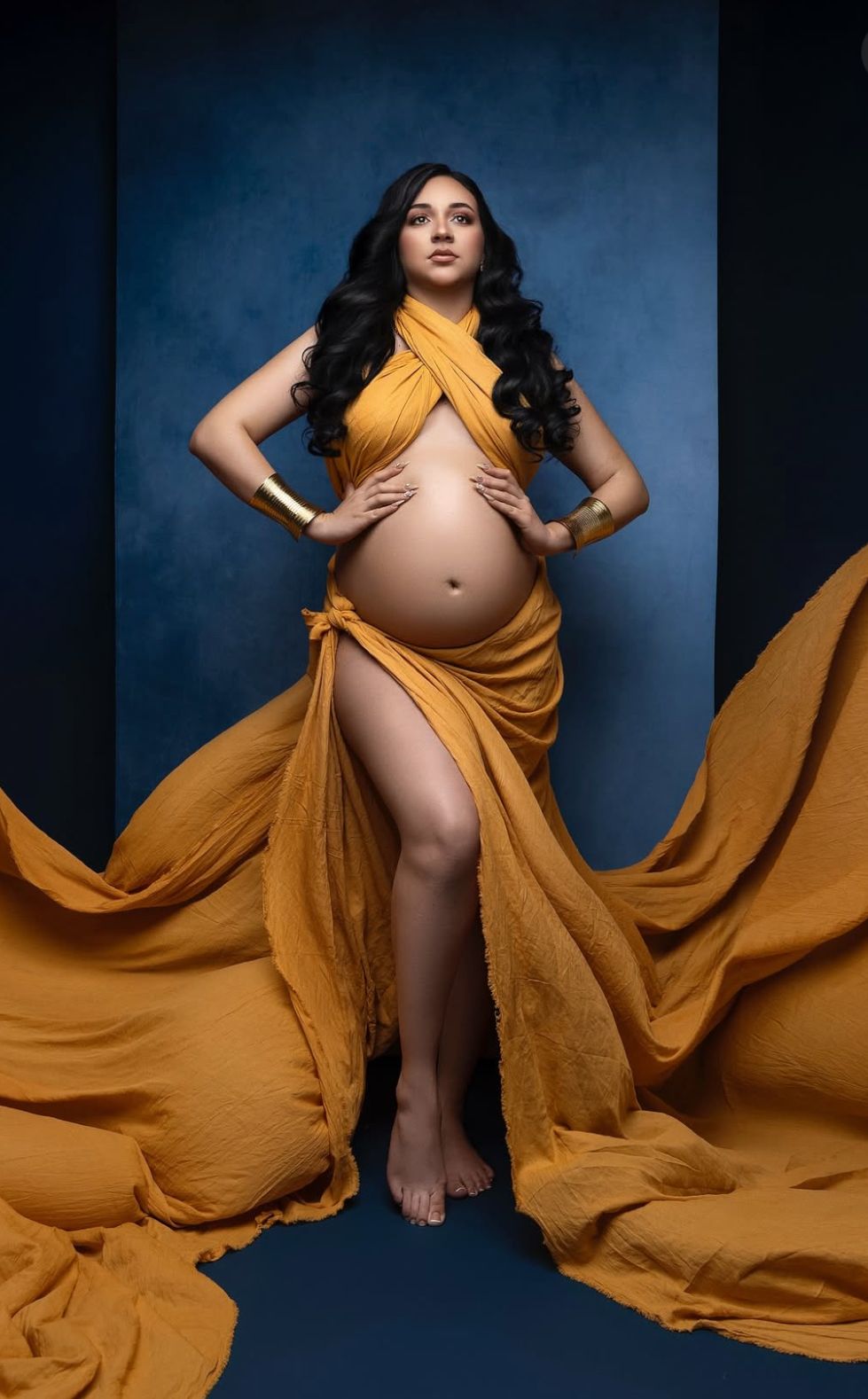 Pregnant woman in flowing golden yellow draped fabric poses gracefully against a blue studio backdrop, showcasing her baby bump.