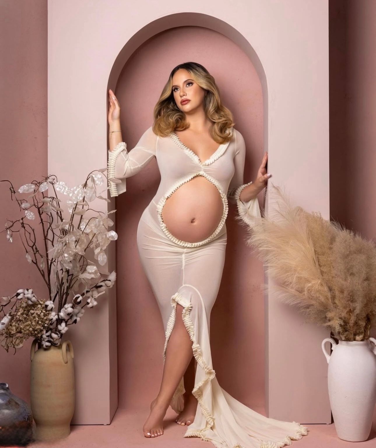Pregnant woman in cream cutout maternity dress posing in pink arch doorway with floral decor.