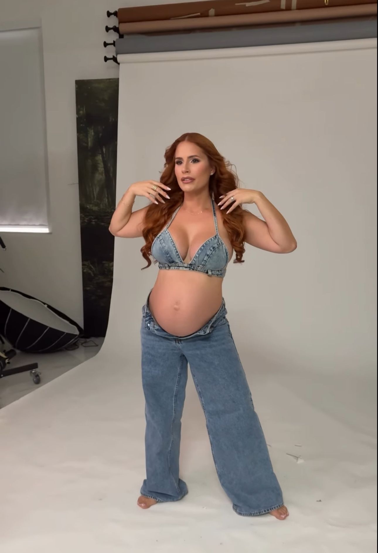 Pregnant woman with red hair poses in denim bralette and wide-leg jeans, showcasing baby bump in a photography studio.