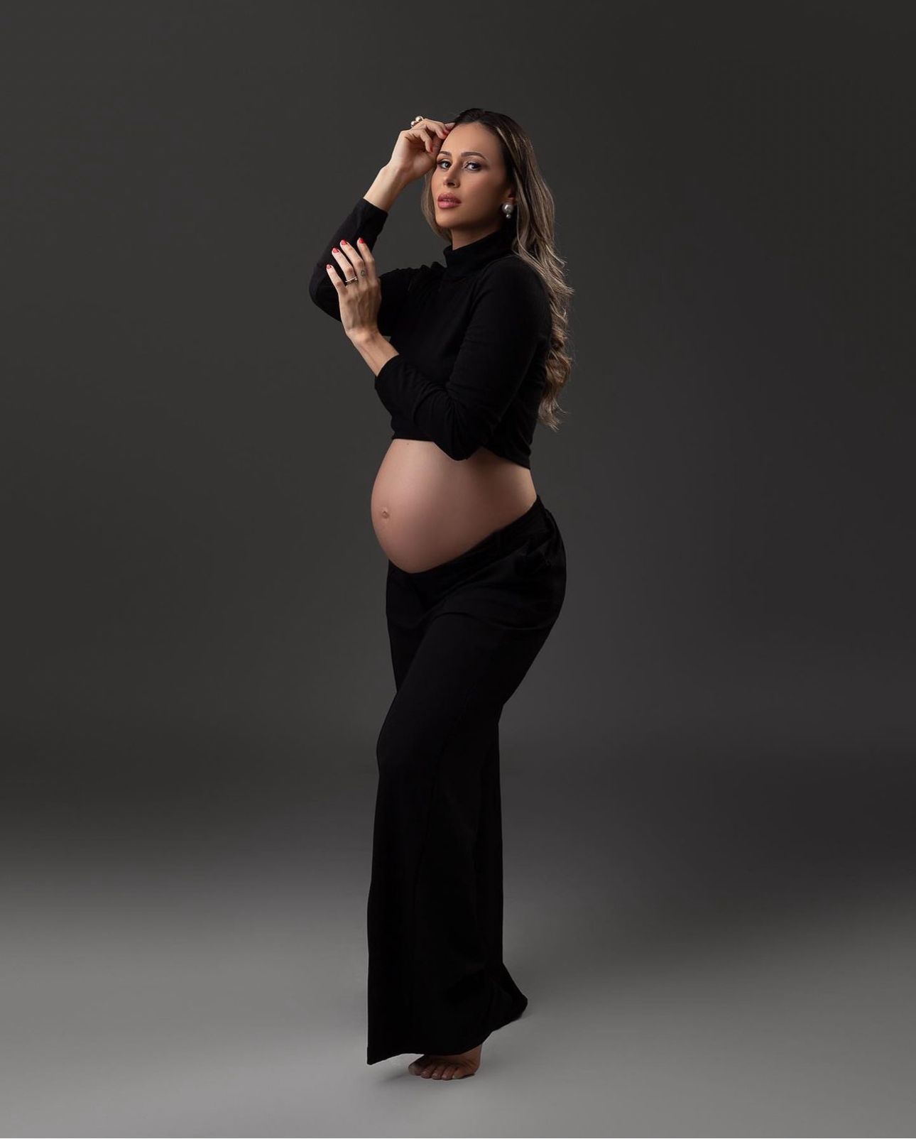 Pregnant woman in black outfit posing for a maternity photo shoot against a gray studio background.