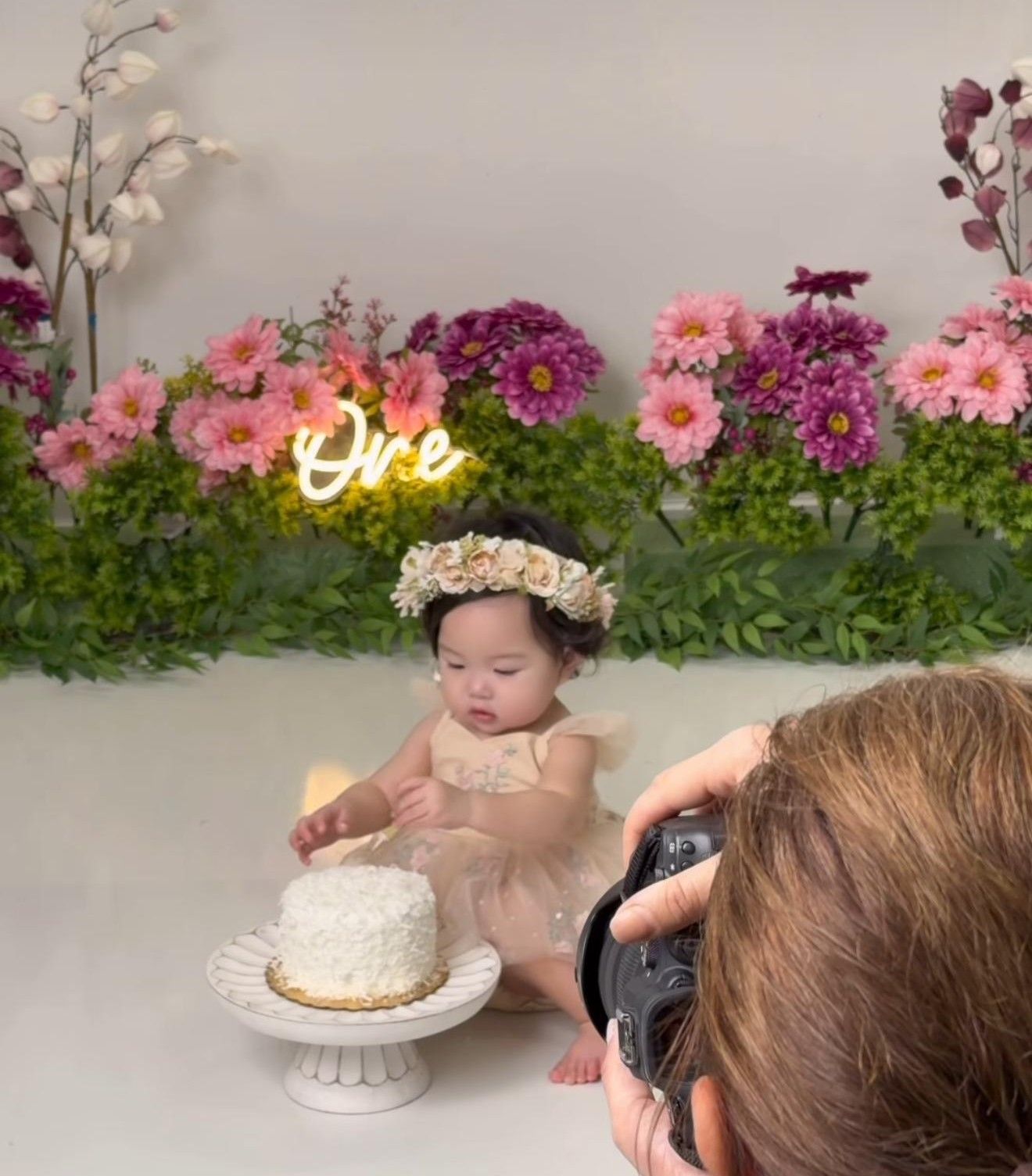 Baby girl in floral dress celebrates first birthday with white smash cake during professional photo shoot.