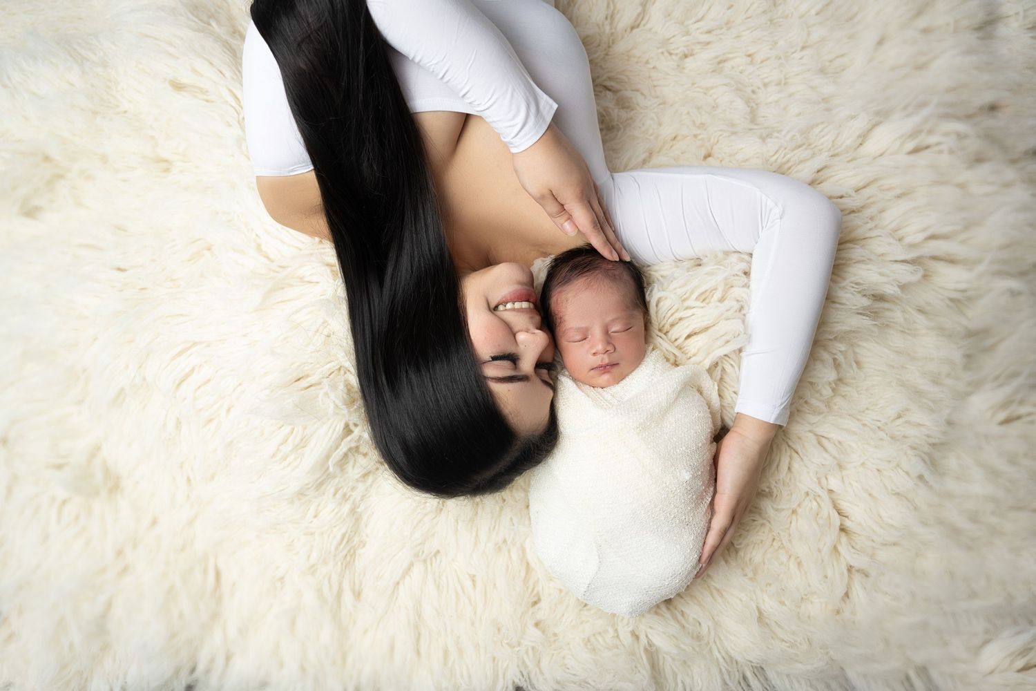 Mother with long dark hair lying on fluffy white rug beside sleeping newborn baby wrapped in white swaddle.