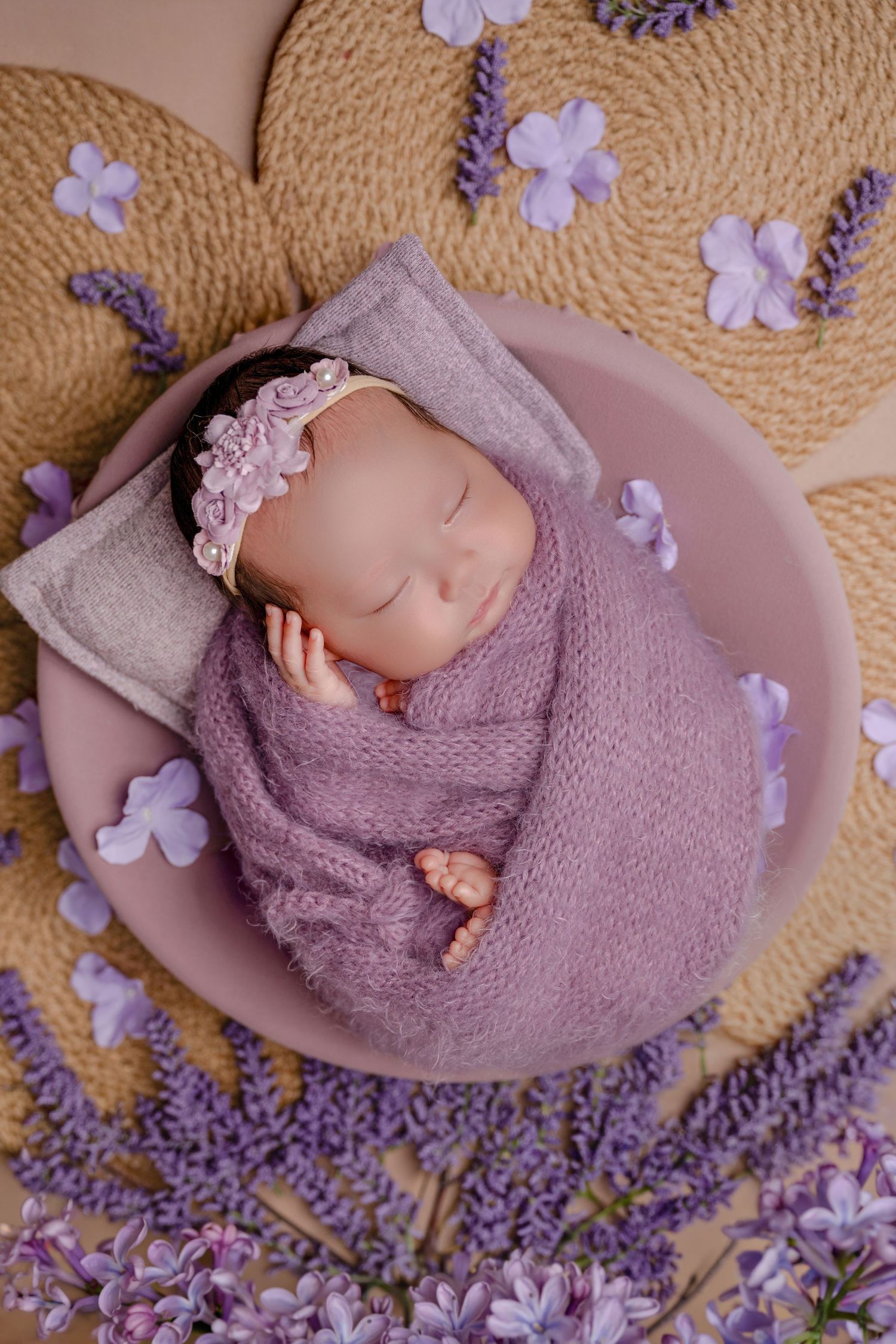Sleeping newborn baby swaddled in purple wrap with floral headband in pink bowl surrounded by lavender flowers.