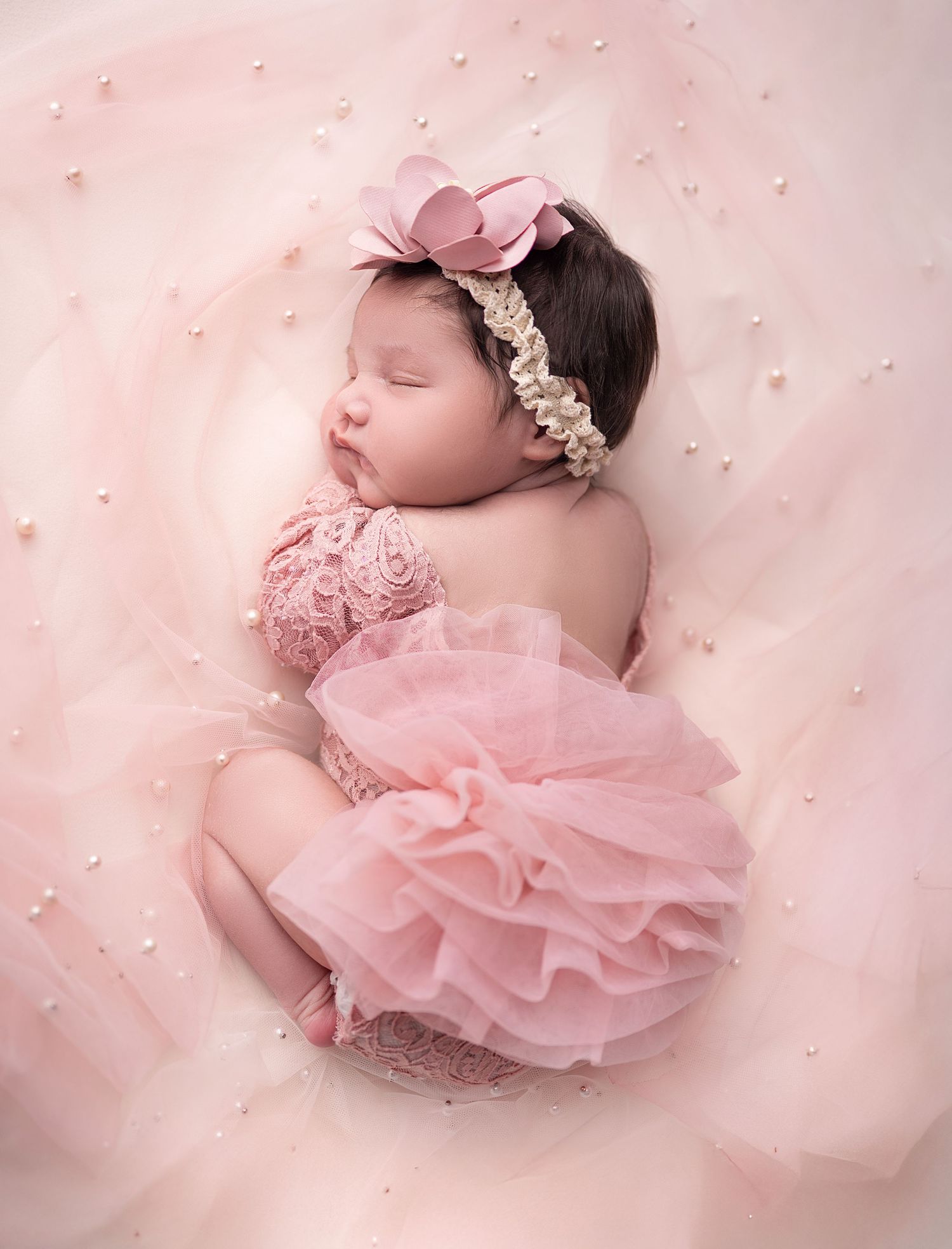Sleeping newborn baby girl in pink tulle dress and floral headband on soft pink pearl-embellished fabric.