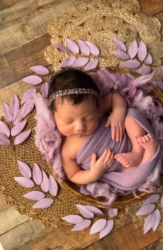 Newborn photography with purple petals and soft fabric in rustic wooden setting.
