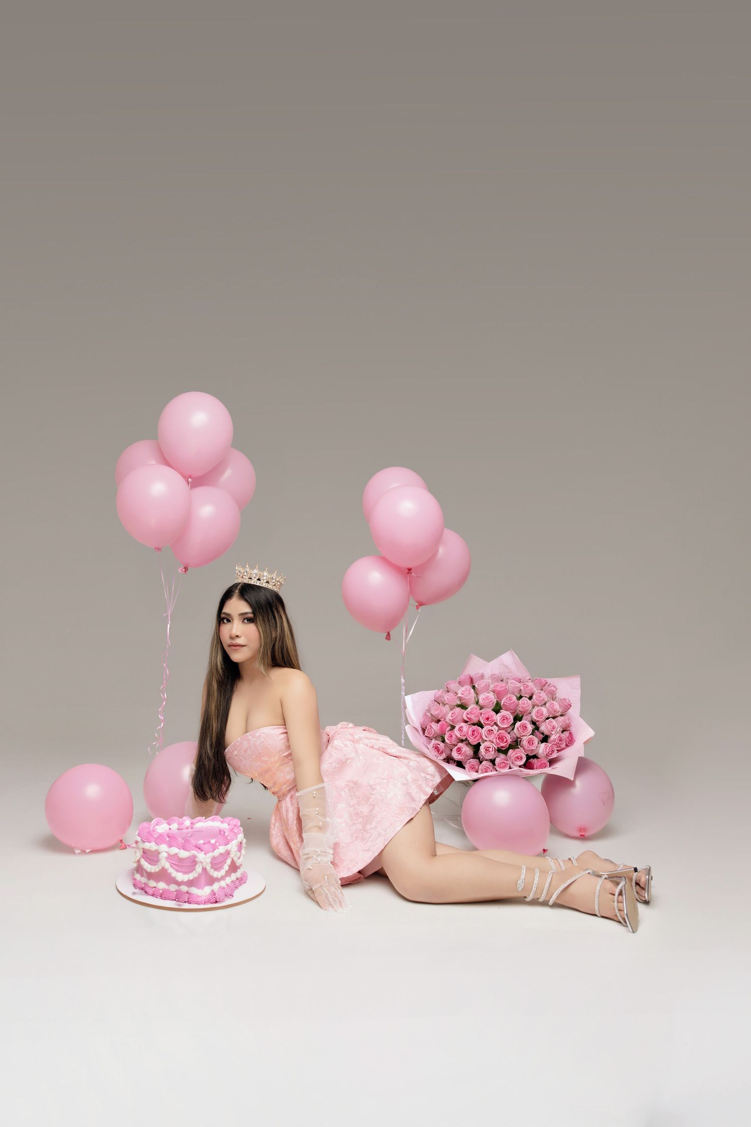Pink birthday celebration setup with balloons, cake, and roses arranged in a minimalist studio setting against grey backdrop.