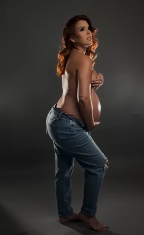 Artistic maternity photo of a pregnant woman in jeans posing against dark background with dramatic lighting.