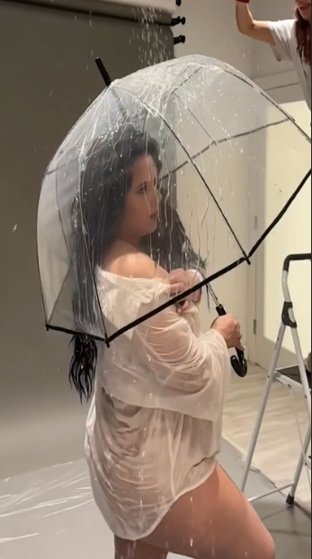 Model poses with clear umbrella in white sheer outfit during studio photoshoot with water effects.