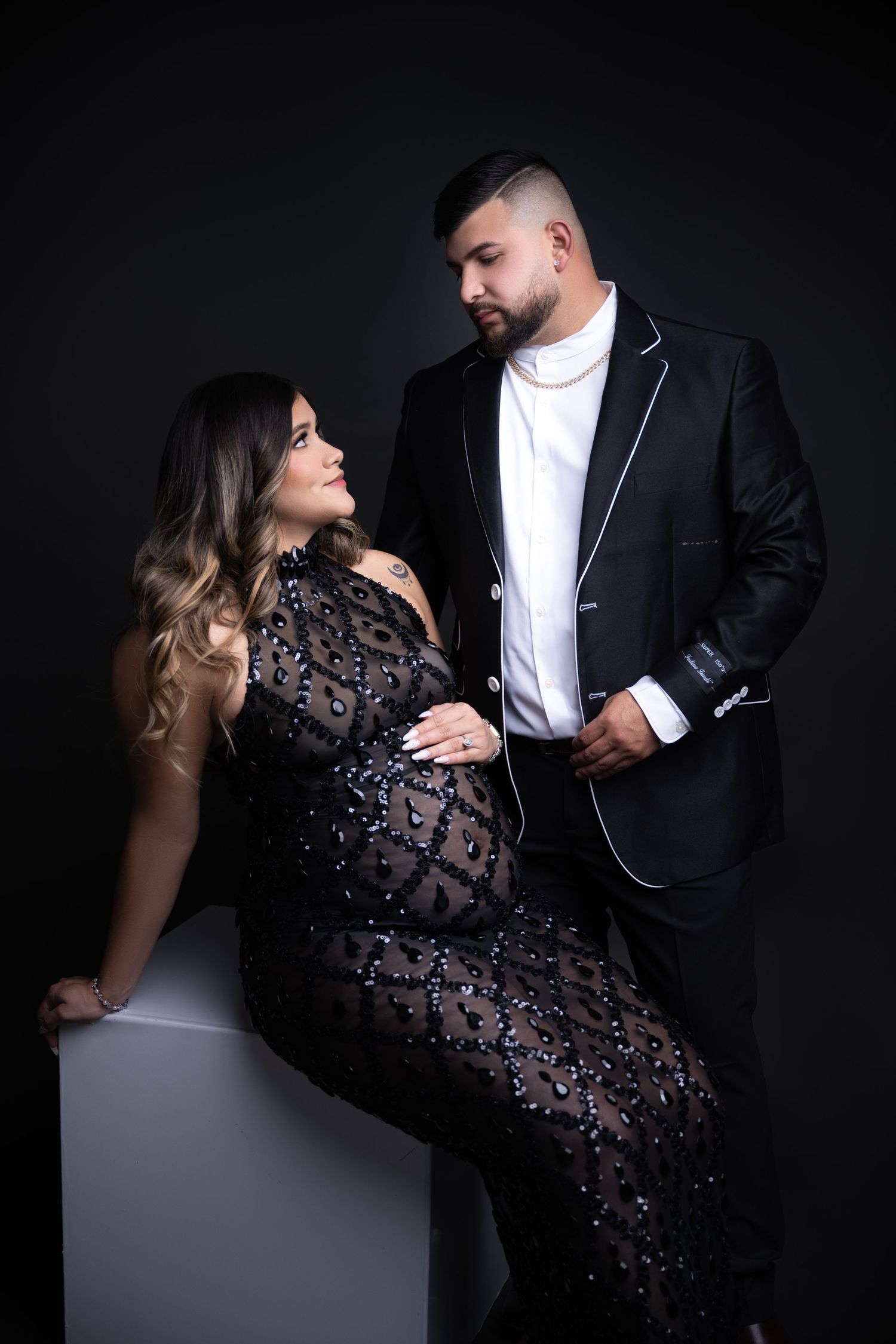 Romantic maternity shoot features couple in formal wear posing against black background.