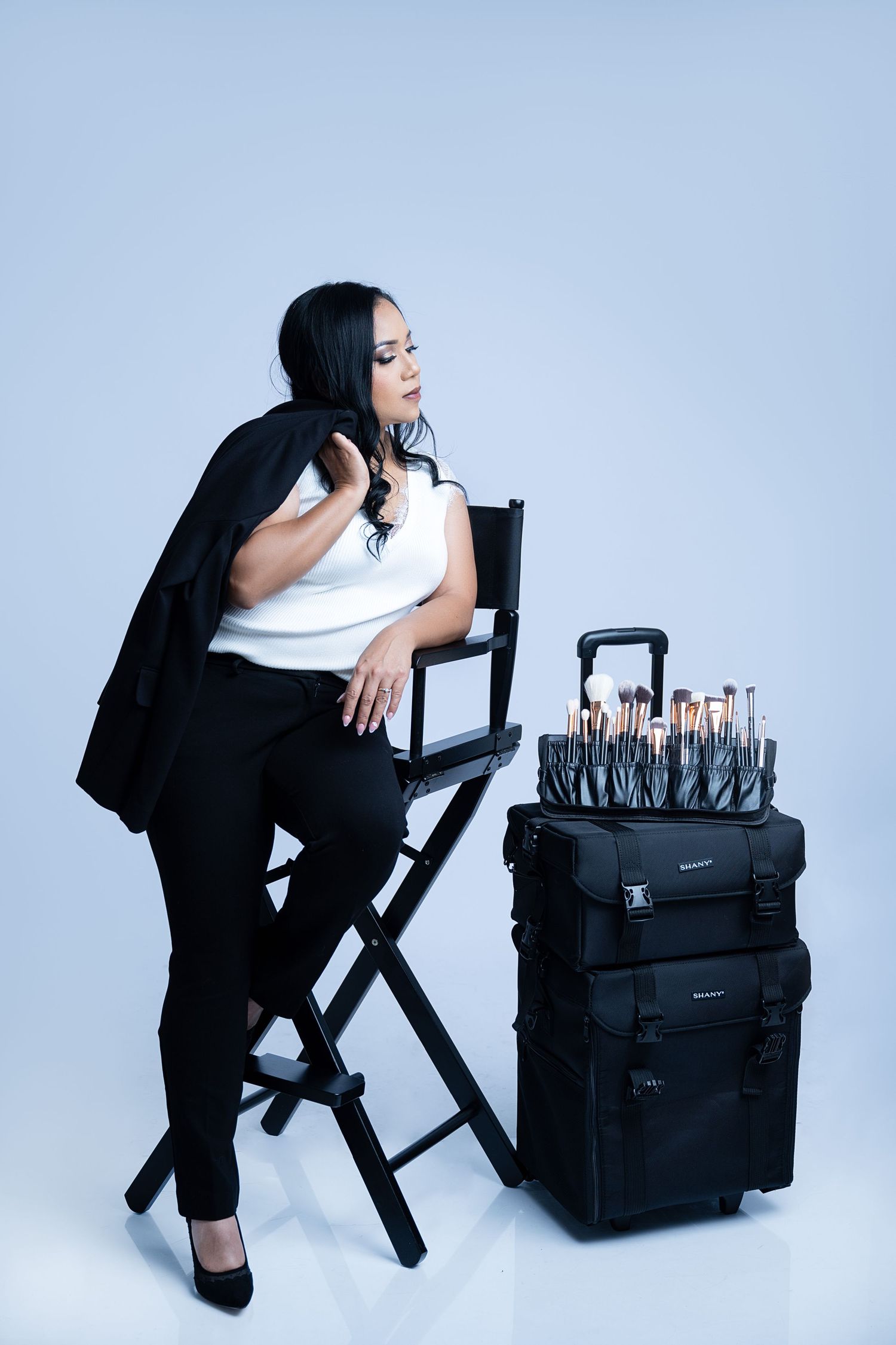 Makeup artist seated in director's chair next to professional brush kit and rolling case.