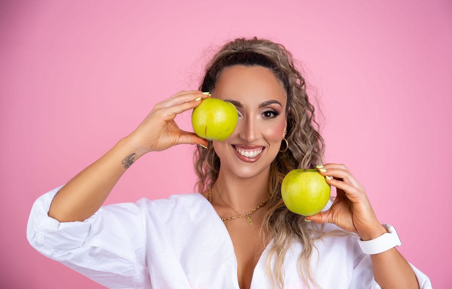 Woman in white blouse holding fresh green apples against pink background with bright smile.