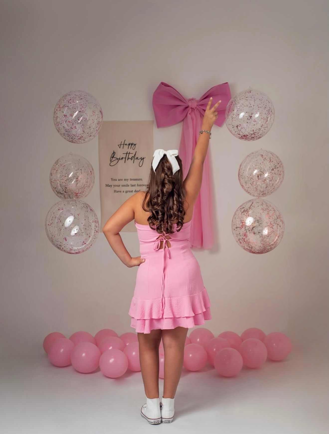 Girl in pink dress reaching for large pink bow on Happy Birthday sign surrounded by pink balloons.