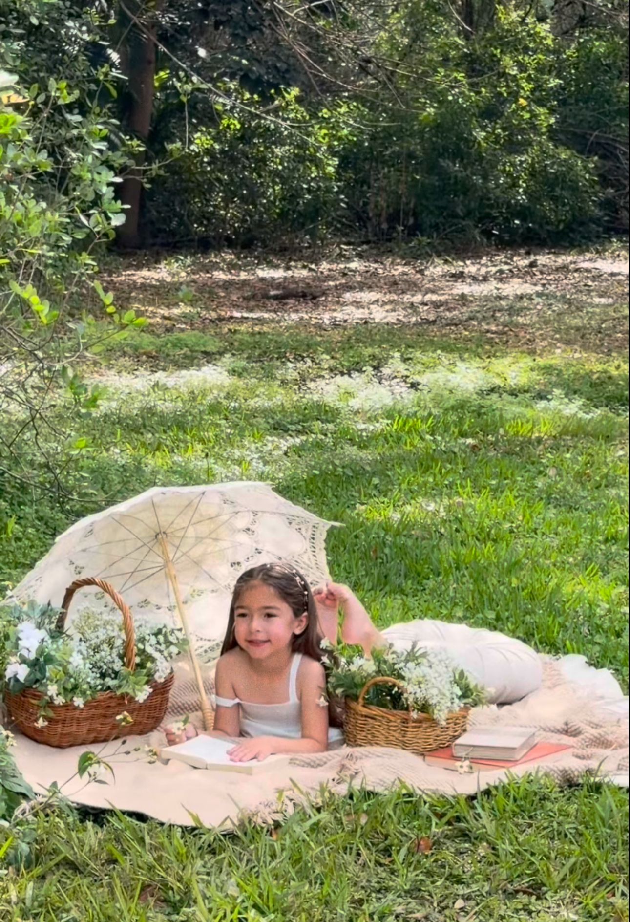 Young girl lying on a blanket outdoors surrounded by flower baskets and a lace parasol during a whimsical garden picnic.