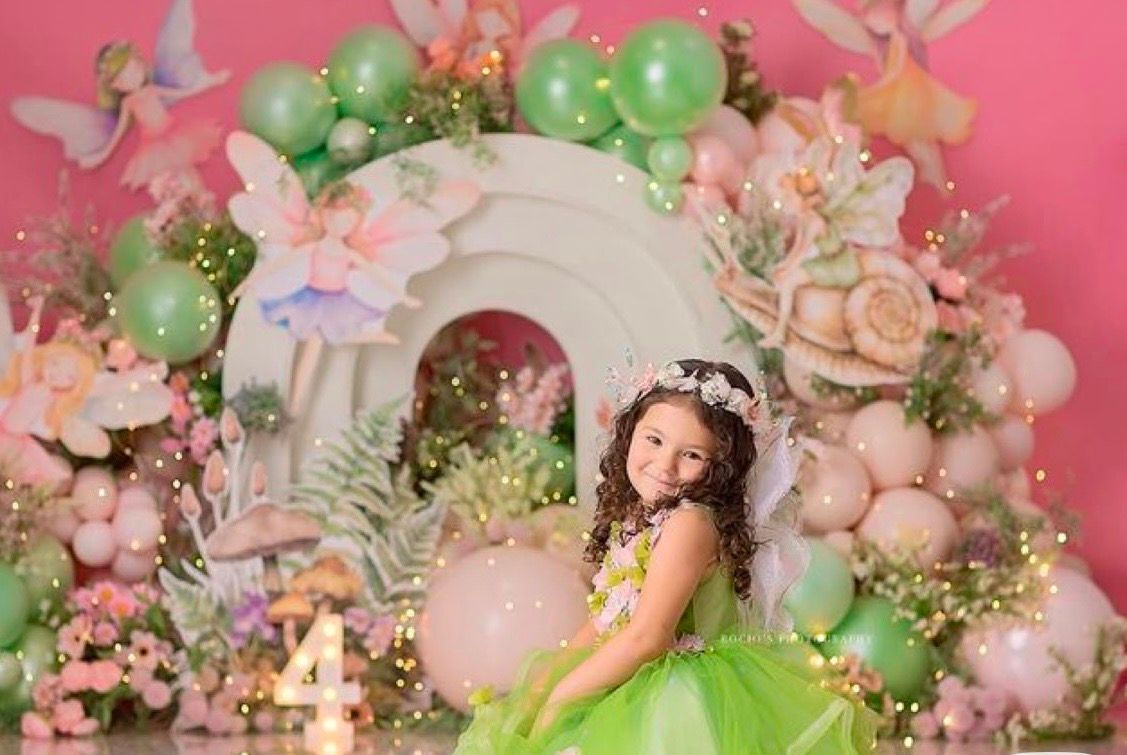 A young girl in a green tutu dress poses in front of a fairy-themed backdrop with pink balloons and flowers.