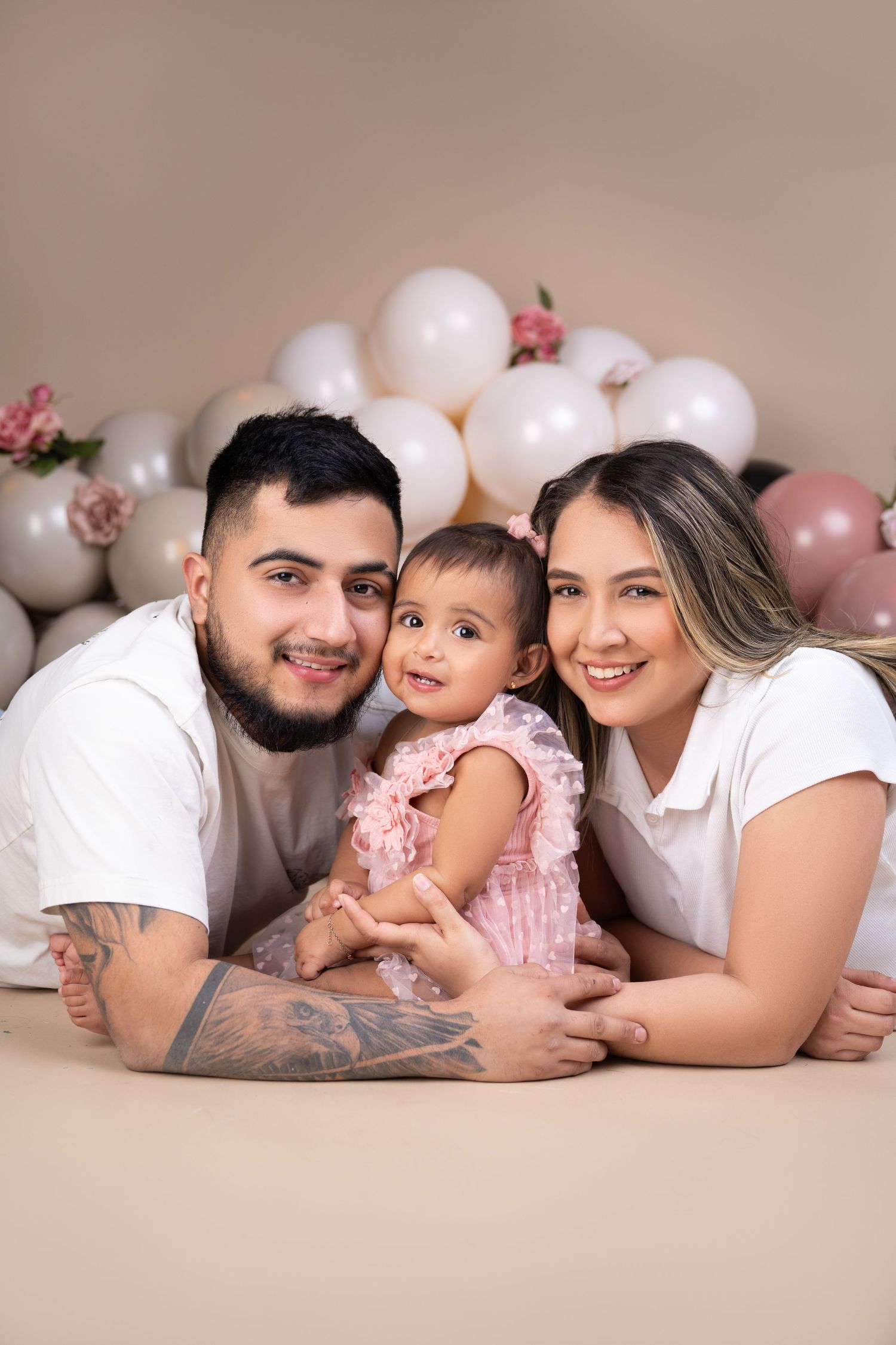 A young family poses with their baby girl in a pink dress, surrounded by white and pink balloons in a studio setting.