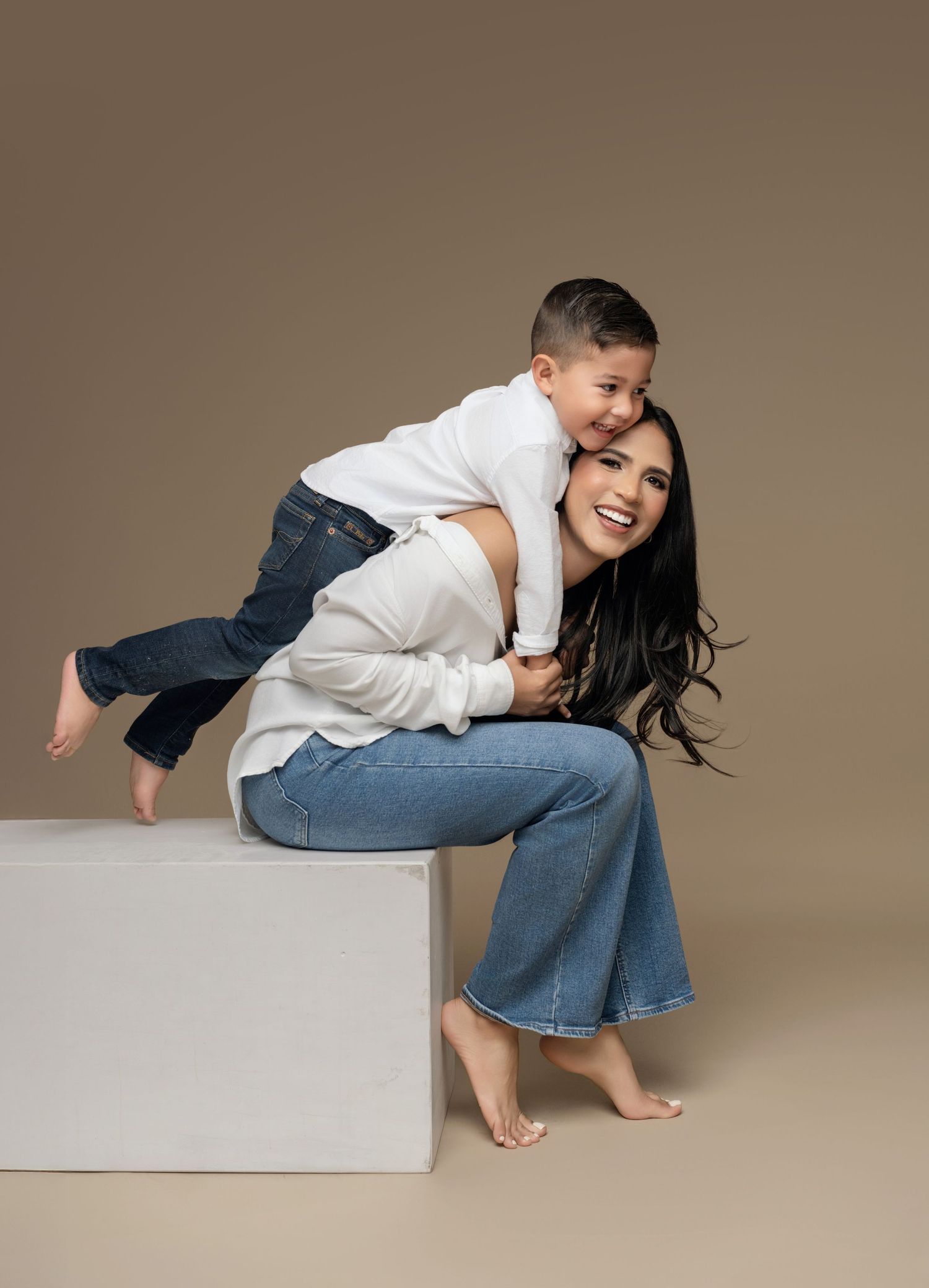 A woman and young boy in matching white tops and jeans pose playfully in a studio, the child hugging her from behind.