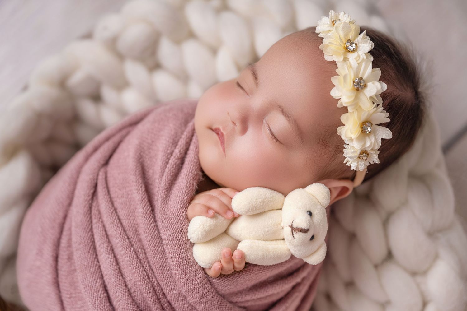 Sleeping newborn baby wrapped in pink swaddle holding tiny teddy bear with floral headband on white knit blanket.