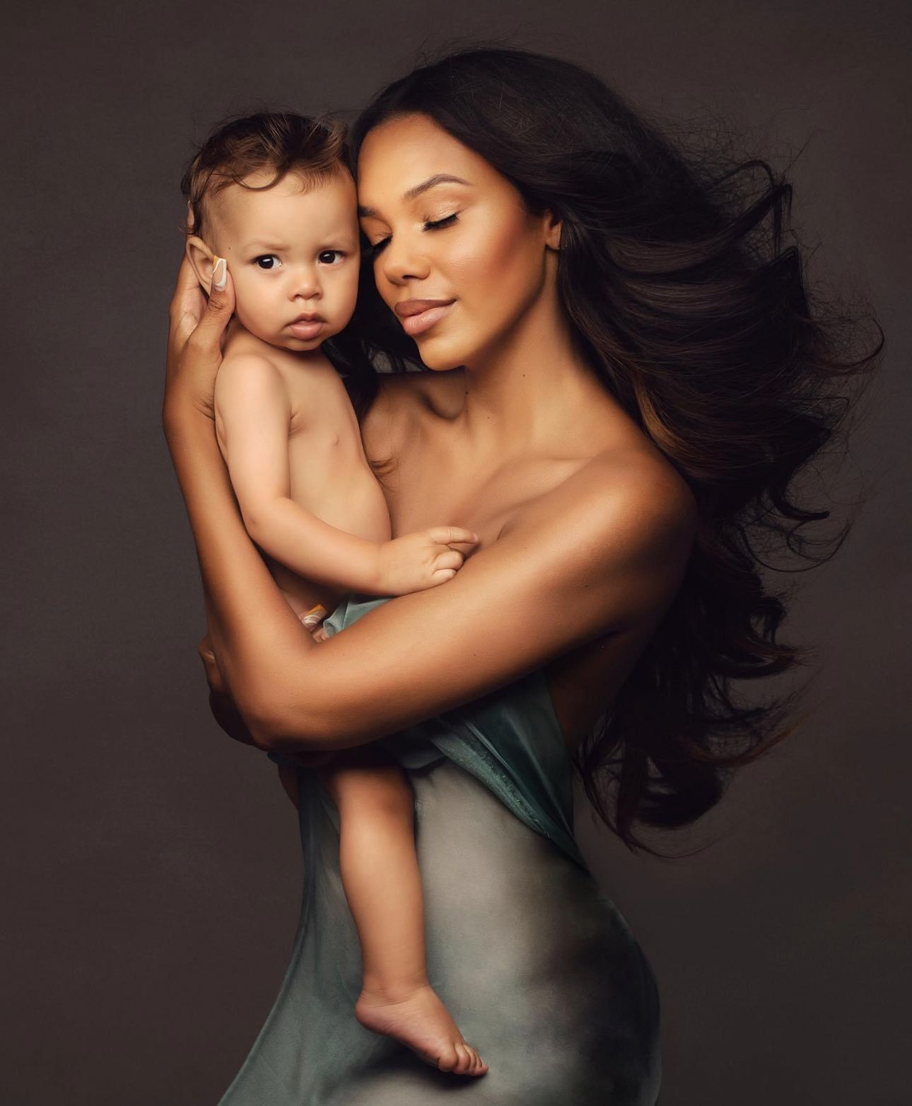 A woman with long dark hair holds a baby tenderly, both posed elegantly against a dark studio background.