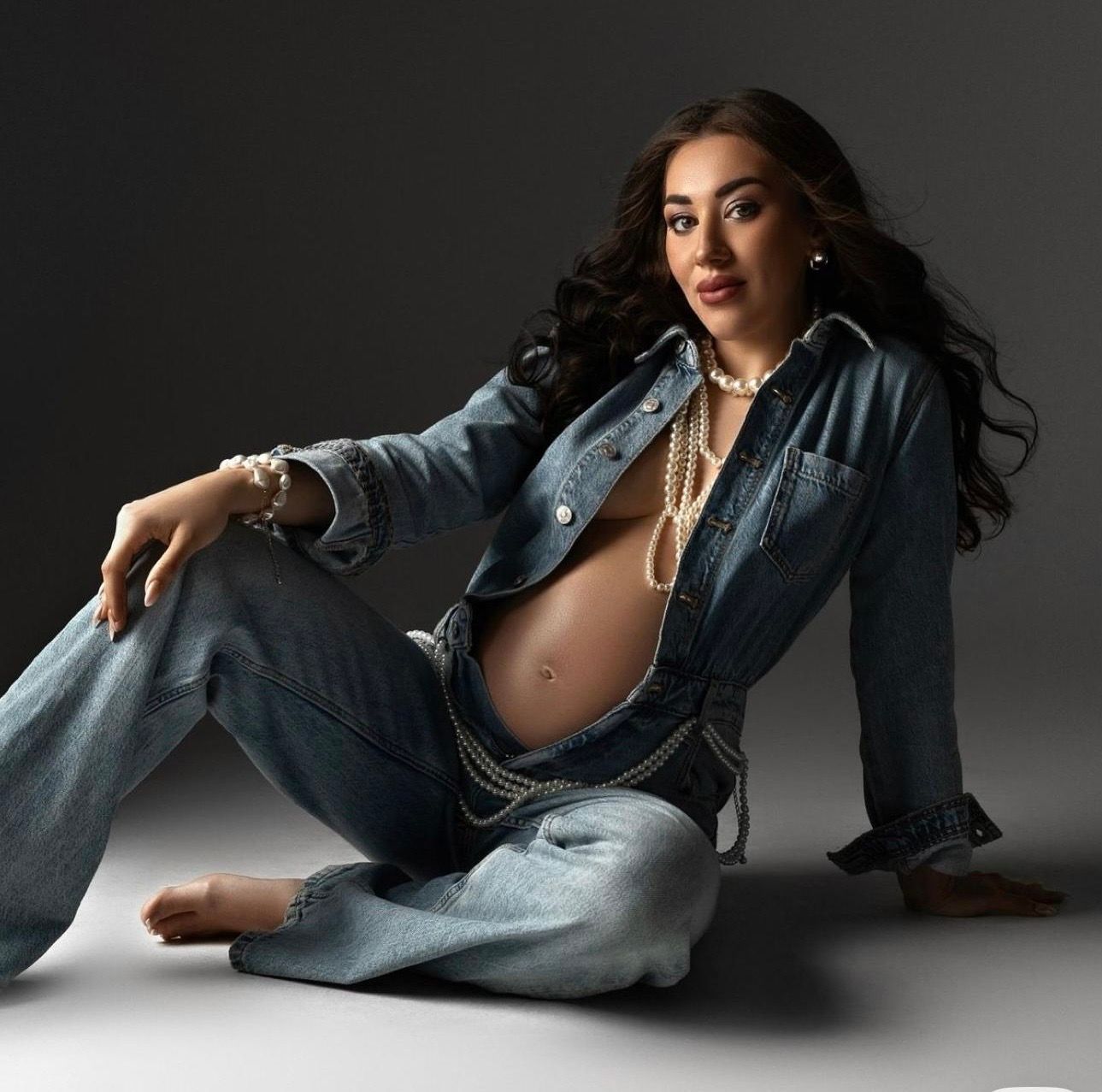 Pregnant woman in denim jacket and jeans with pearl necklace, sitting gracefully on studio floor.