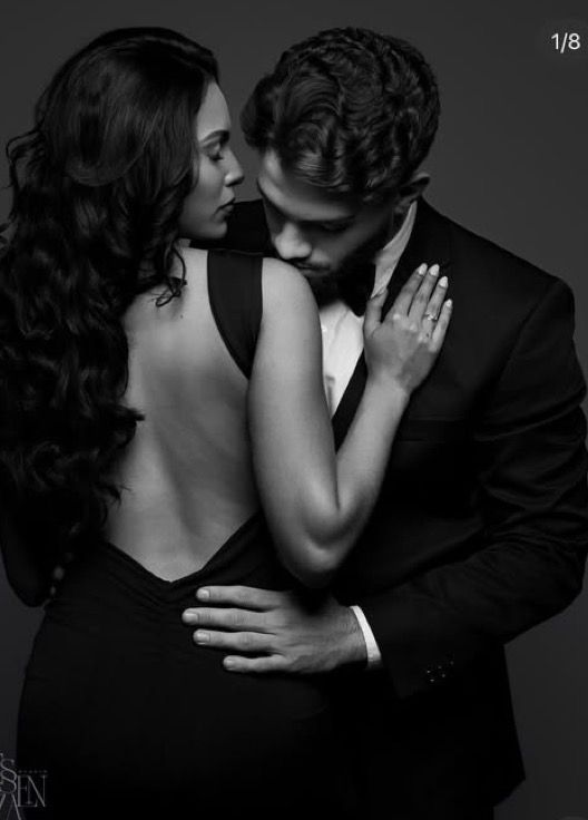 Elegant black and white couple portrait, woman in backless gown embraced by man in tuxedo in romantic pose.