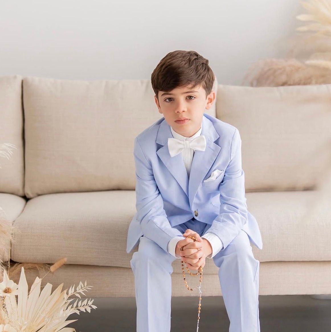 A young boy in a light blue suit with white bow tie sits on a beige sofa holding rosary beads.