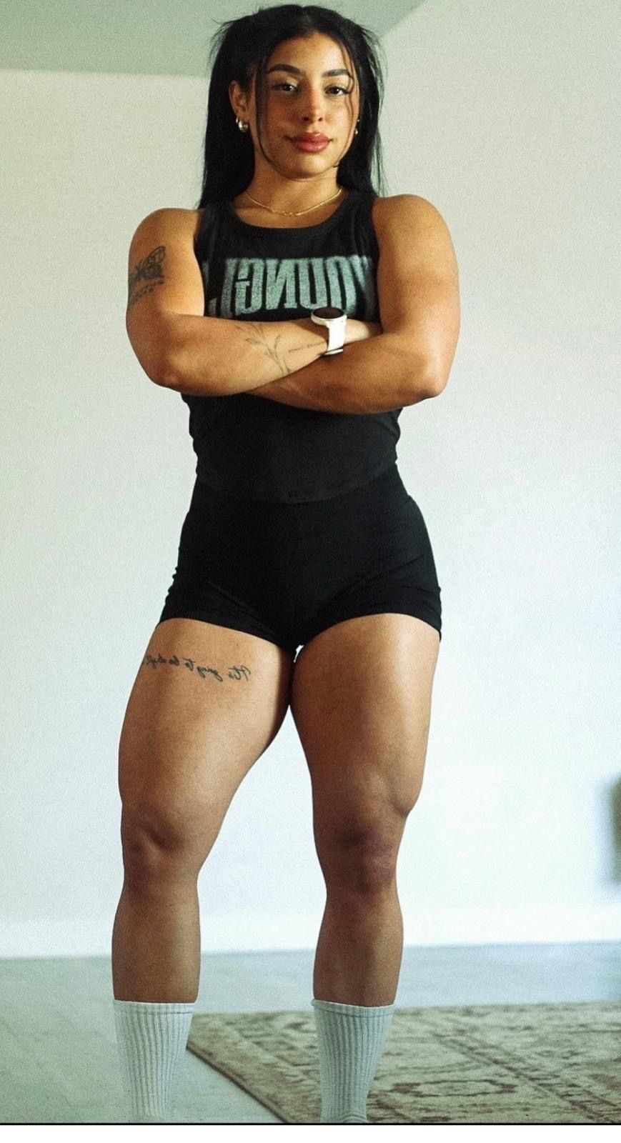 Muscular woman in black YoungLA tank and shorts stands with arms crossed showing defined legs.