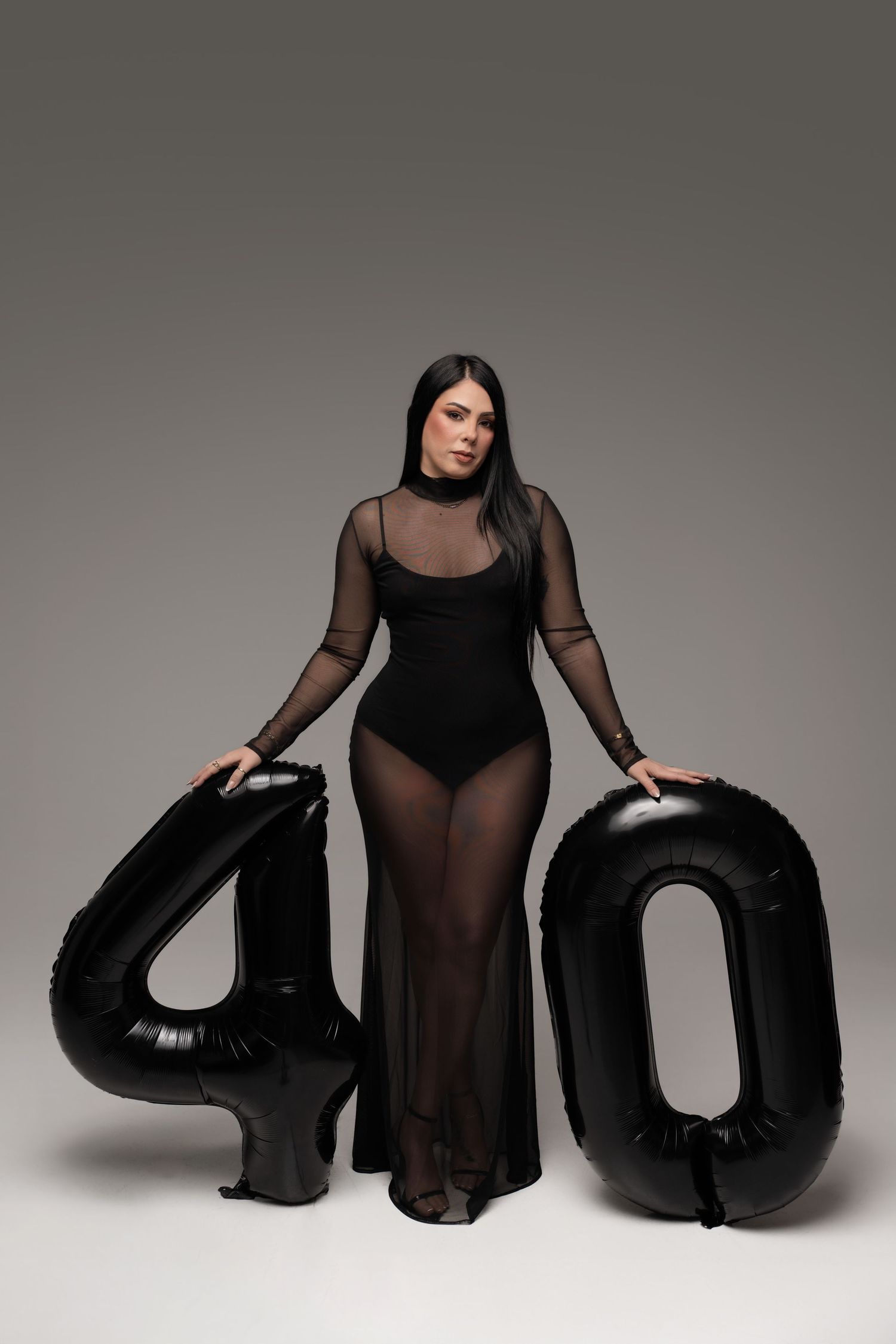 Woman in black mesh bodysuit posing with large black number 40 balloons on gray background.