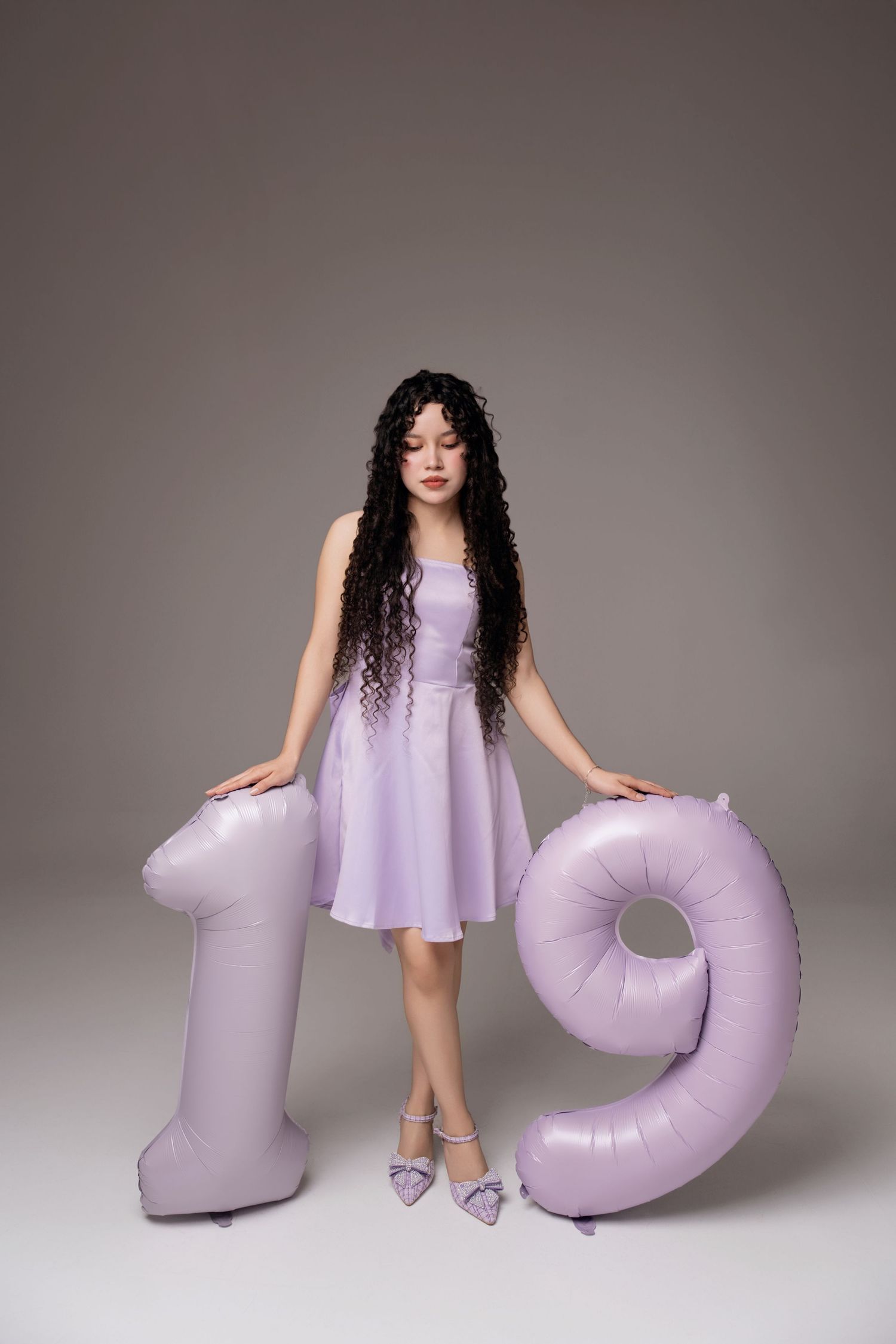 Young woman in purple dress posing with large number 19 balloons on gray background.