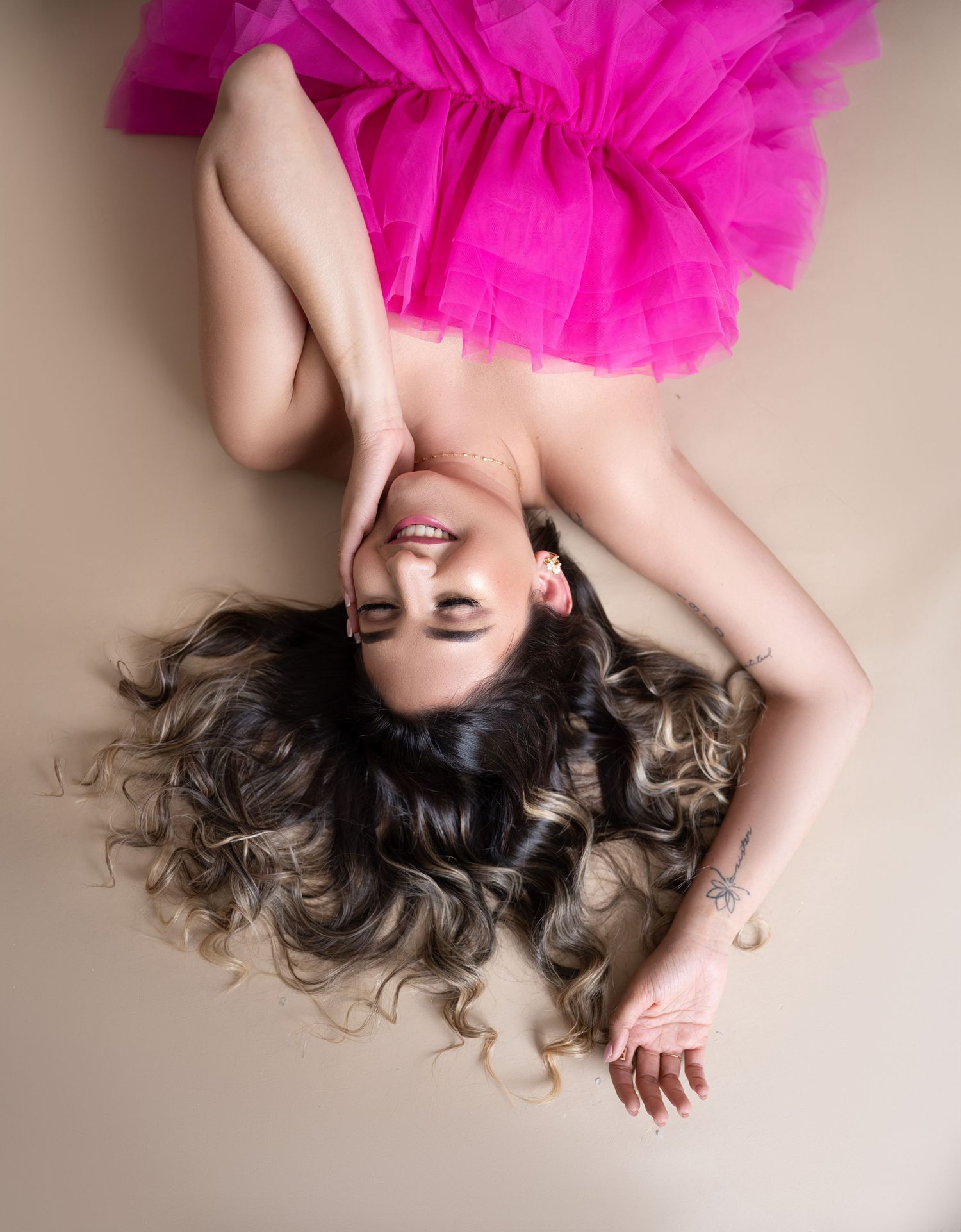 Woman lying on a beige backdrop in a bright pink tulle dress, smiling with eyes closed and long wavy hair fanned out.