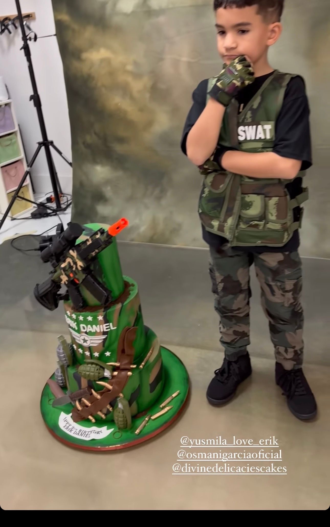 Young boy in a SWAT vest and camouflage outfit stands beside a green military-themed tiered birthday cake topped with a toy rifle.