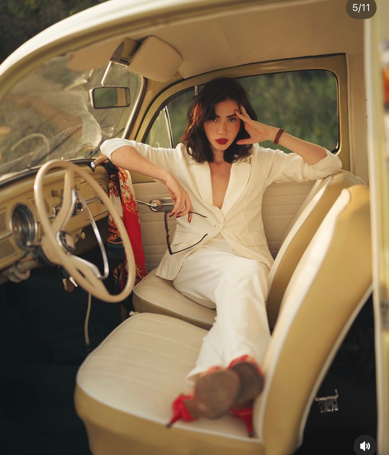 Woman in white suit posing in vintage cream car interior, red lips, dark hair, holding handbag.