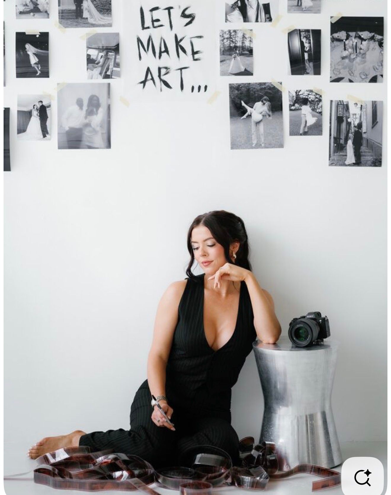 A woman in a black outfit sits on the floor beside a camera, surrounded by film strips, with art photos on the wall.