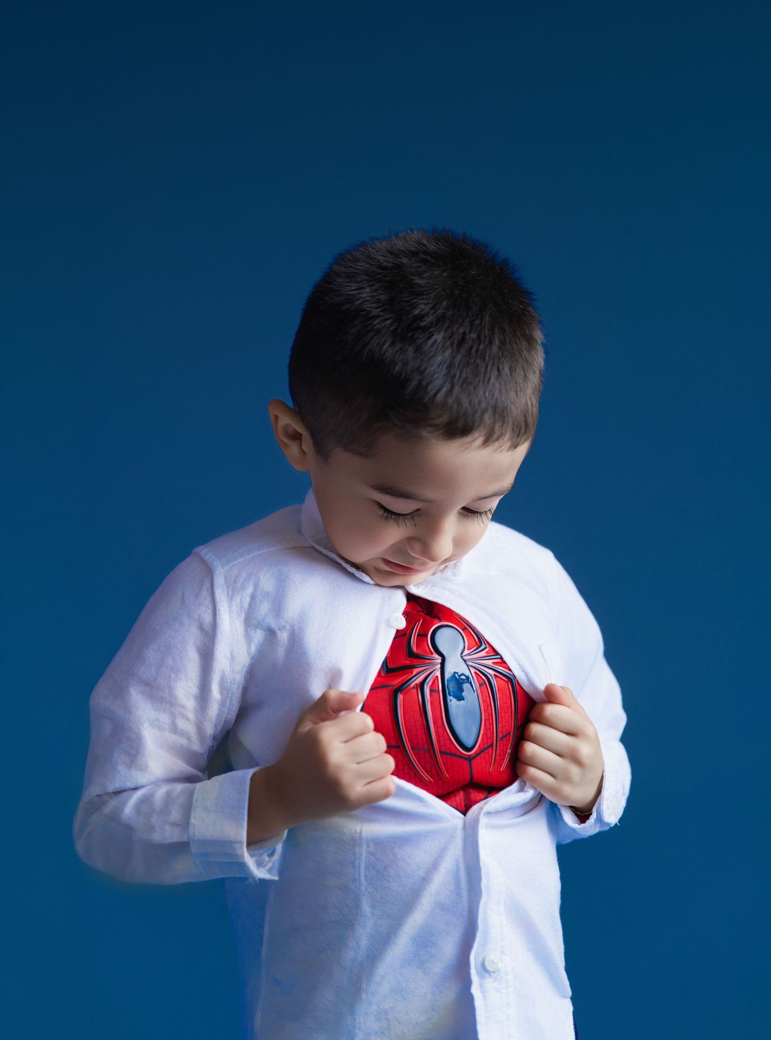 Young boy opening white shirt to reveal Spider-Man costume underneath, posing heroically against blue background.