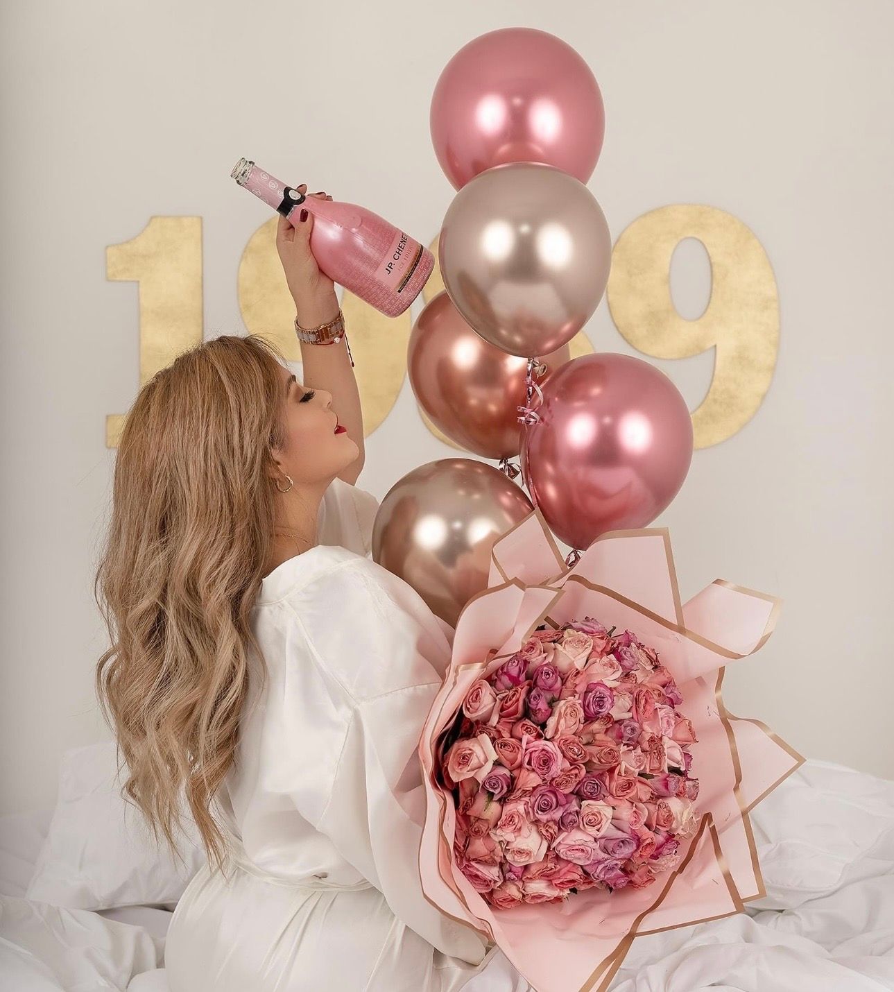 Woman holding pink champagne bottle with rose gold balloons and large pink flower bouquet near gold number decorations.