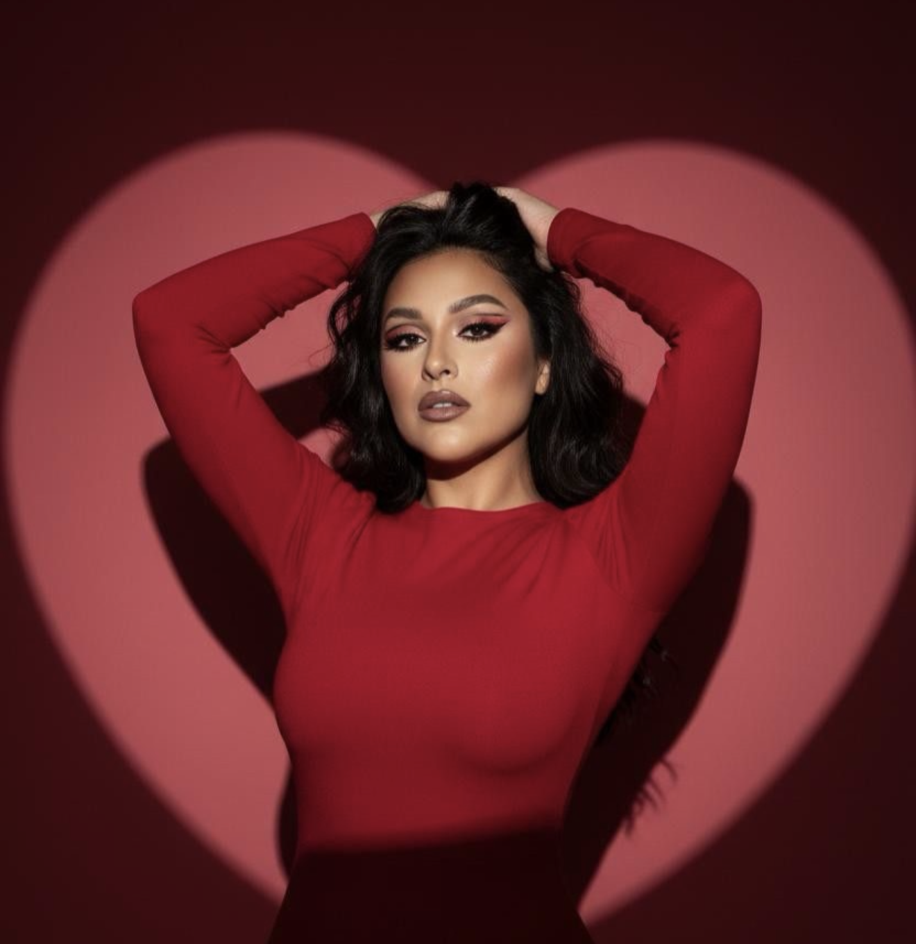 Woman in red long-sleeve top poses against heart-shaped pink spotlight on red background.