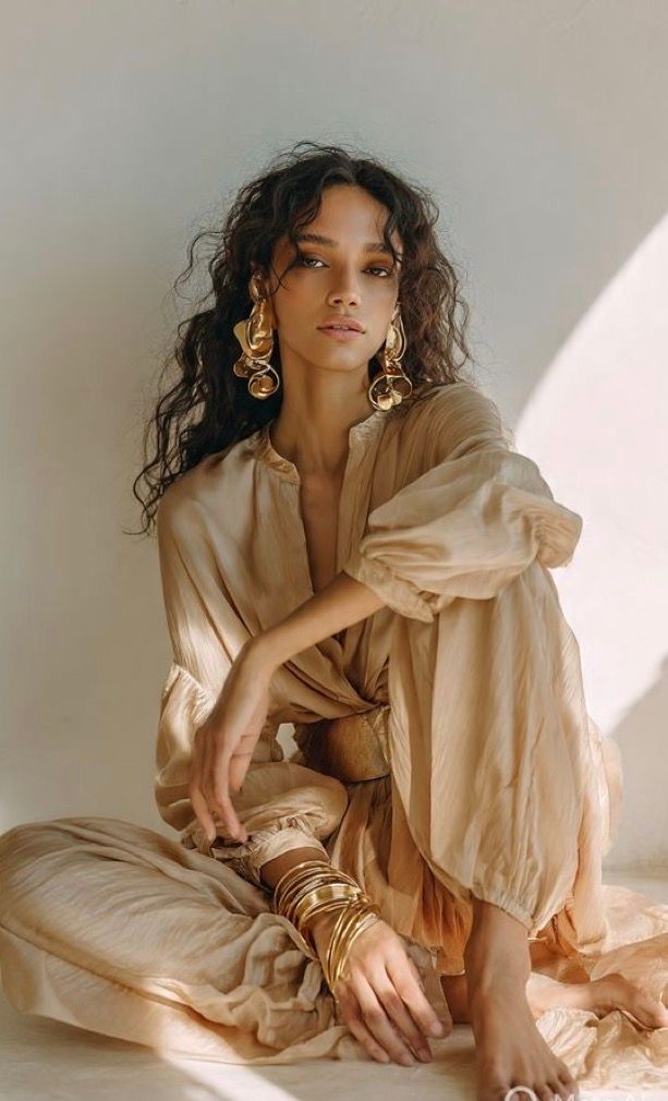 Woman in flowing beige linen outfit with gold bangles and statement earrings, seated gracefully on floor.